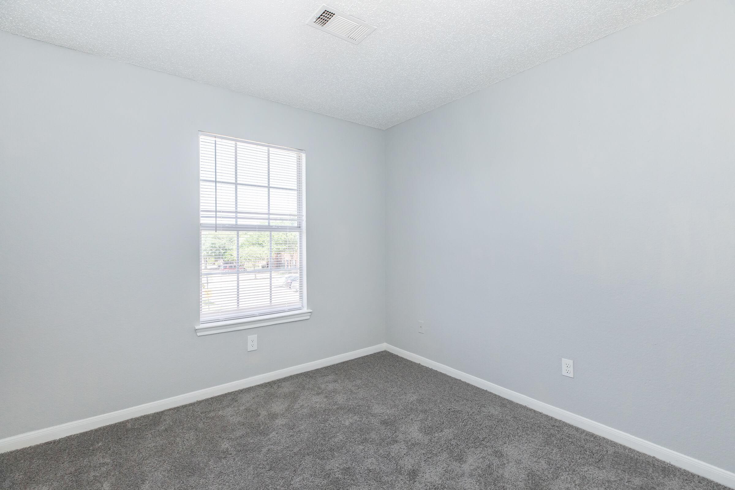 Empty room with light gray walls and carpet. A single window with blinds allows natural light to enter. The room has a simple design with no furniture or decorations, providing a blank canvas for potential personalization.