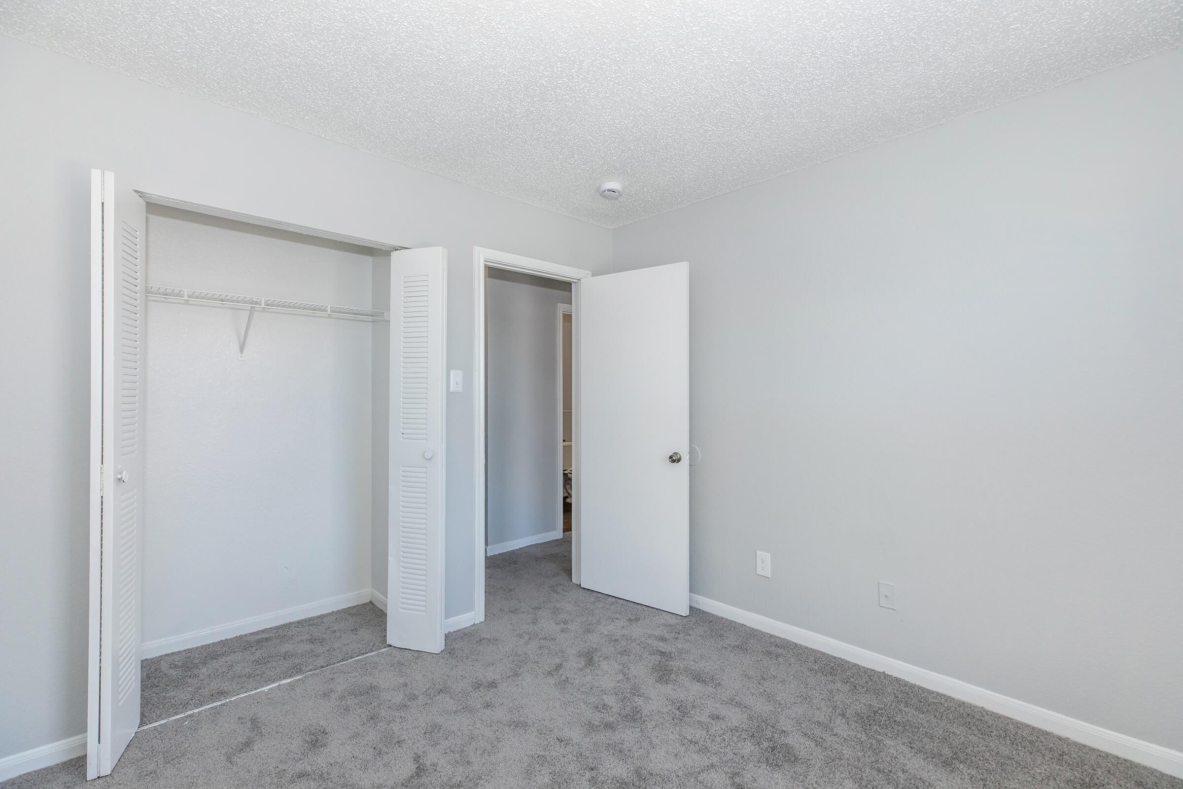 A neutral-colored bedroom with light gray walls and carpet. The room features two open doors leading to closets, one with shelving, and a doorway to another space. Natural light illuminates the area, creating a bright and spacious feel.