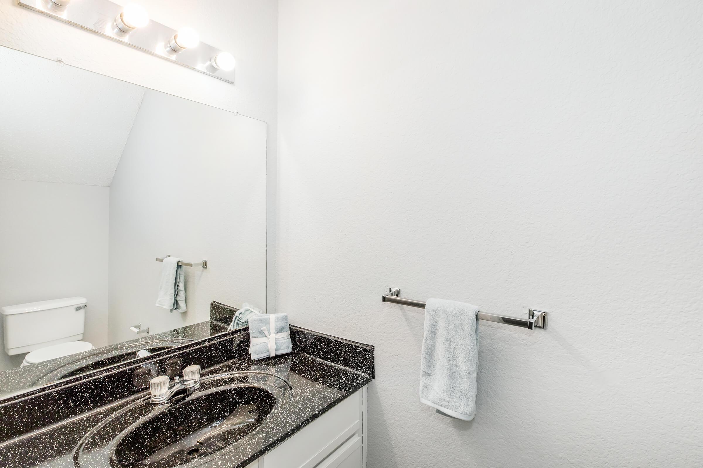 Bathroom interior featuring a black countertop, a sink with two faucets, a polished mirror above the sink, and a towel hanging on a silver rack. In the background, a white toilet is visible, and the walls are painted in a light shade. Soft lighting is provided by a fixture above the mirror.