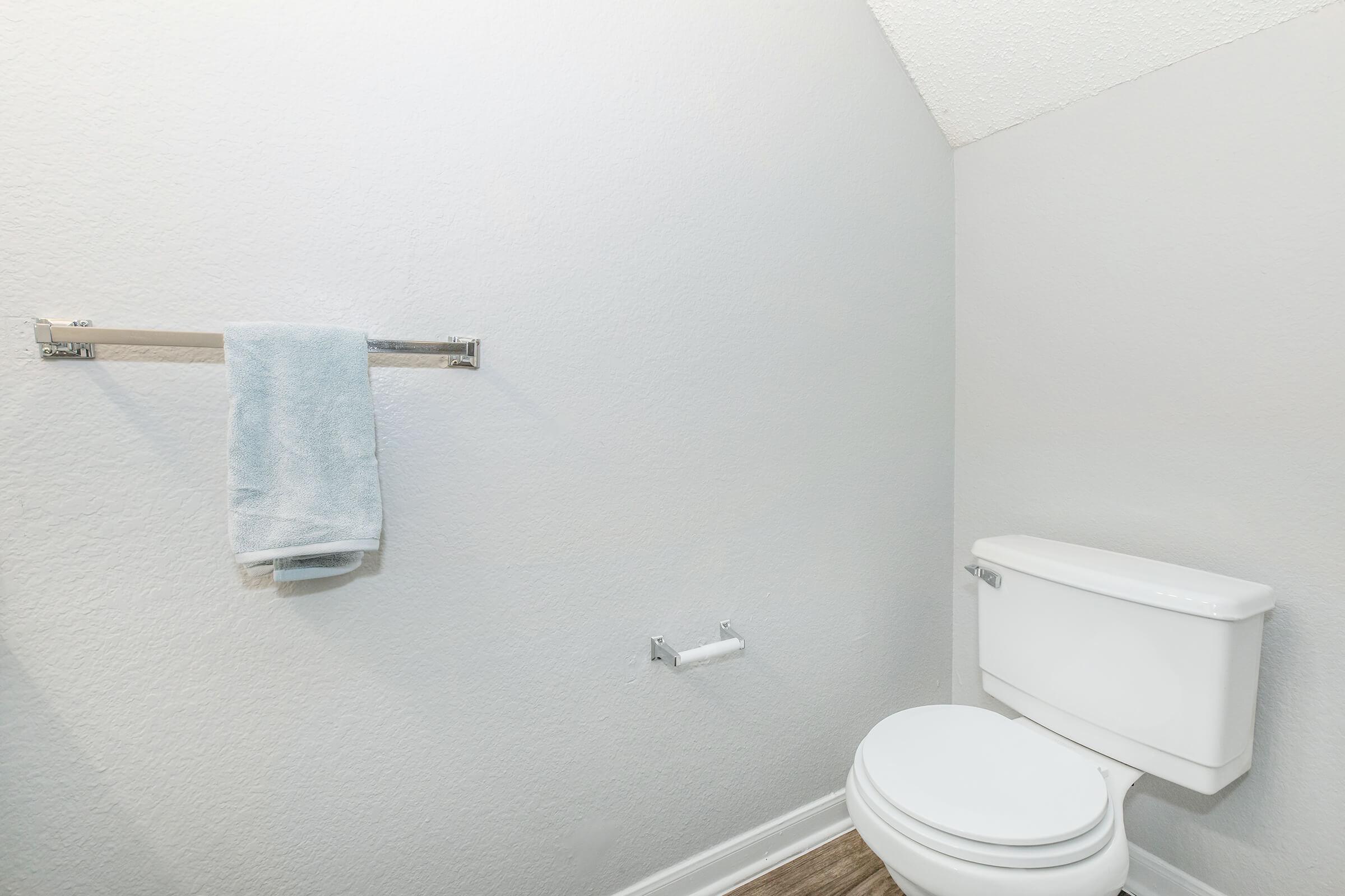A small bathroom with light gray walls. There's a white toilet and a toilet paper holder next to it. A silver towel rack holds a light blue towel, and the floor is covered with light brown flooring. The overall space is tidy and simple.