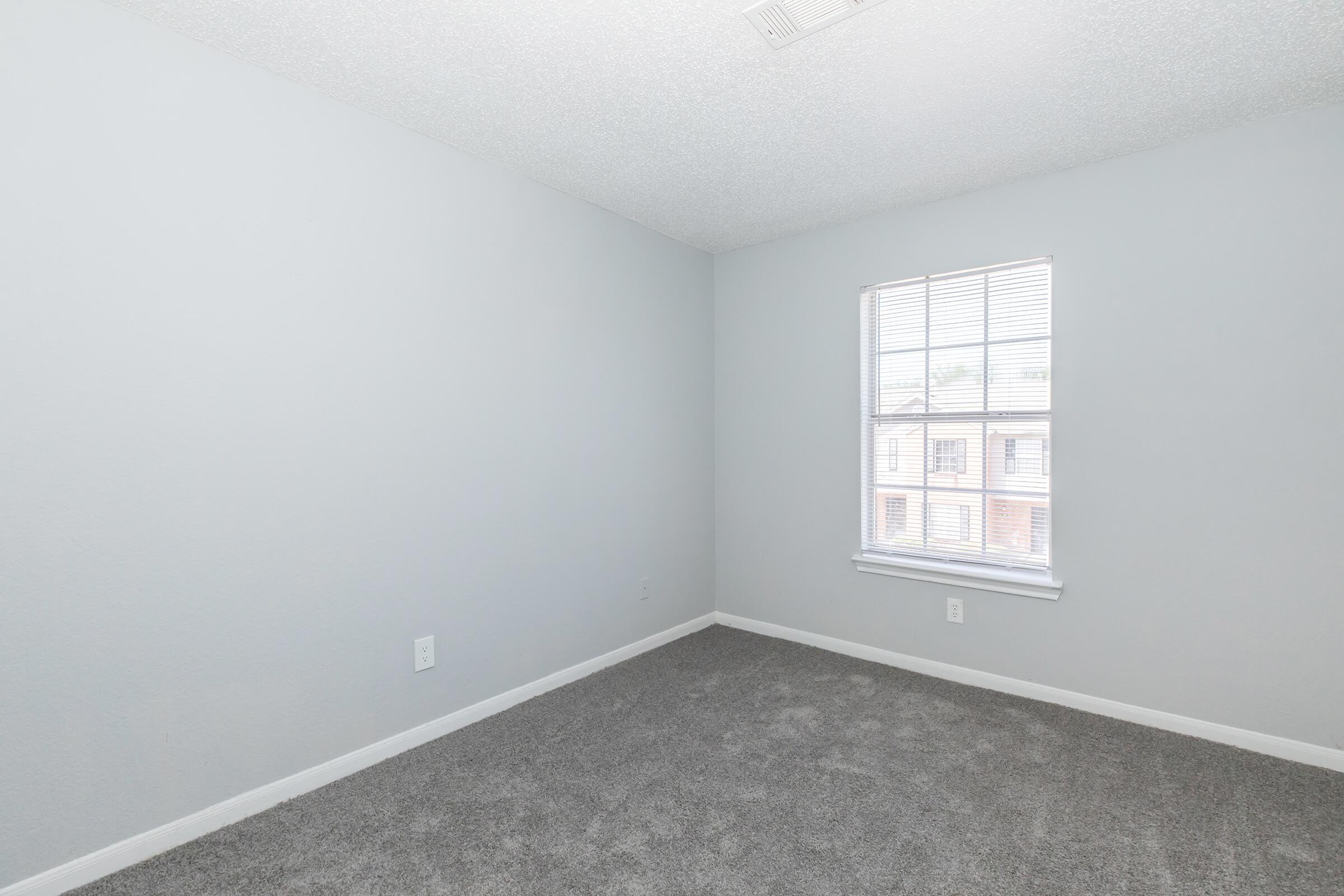 A lightly colored room with soft gray walls and plush gray carpet. A single window with blinds allows natural light to enter, illuminating the space. The room appears empty and is suitable for various uses, such as a bedroom or office. No furniture is present, creating a minimalist atmosphere.