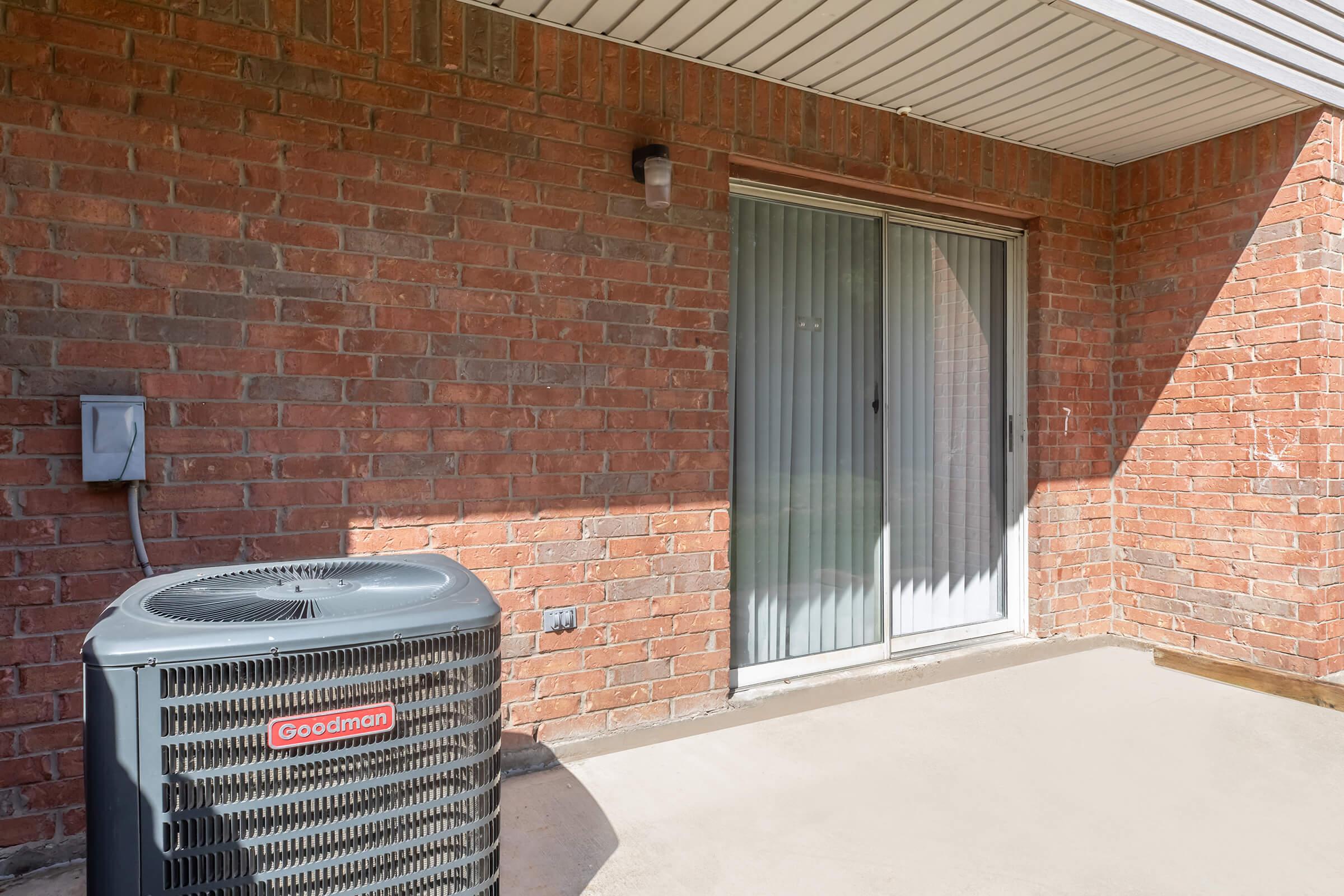 View of a patio area featuring a sliding glass door with sheer curtains and a brick wall. An air conditioning unit is positioned to the left. The floor is concrete, and the scene is well-lit with shadows cast from the structure above.