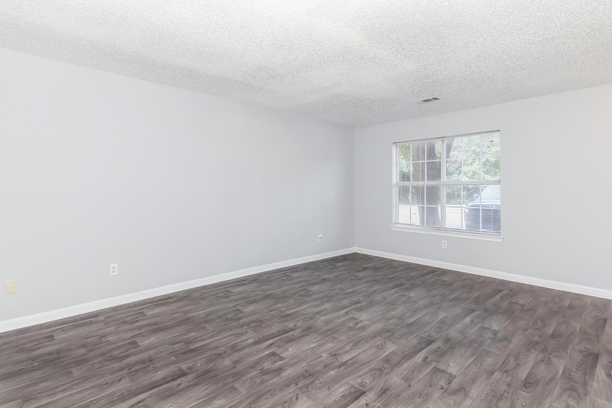 A spacious, empty room with light gray walls and a textured ceiling. The floor features dark wood-like laminate. A large window allows natural light to enter, providing a view of greenery outside. There are no furnishings, creating a blank canvas for potential decoration.