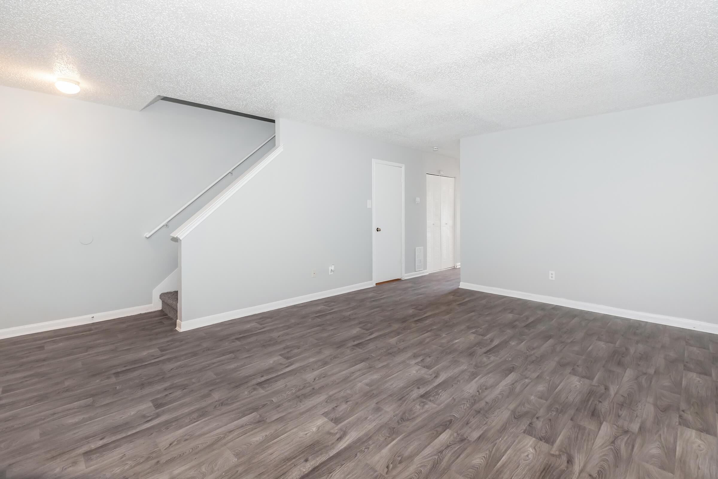 An empty living room featuring light gray walls and a new wood-look floor. A staircase is visible on the left side, with a white door and light coming from a nearby window in the background. The space is clean and unfurnished, creating a bright and open atmosphere.