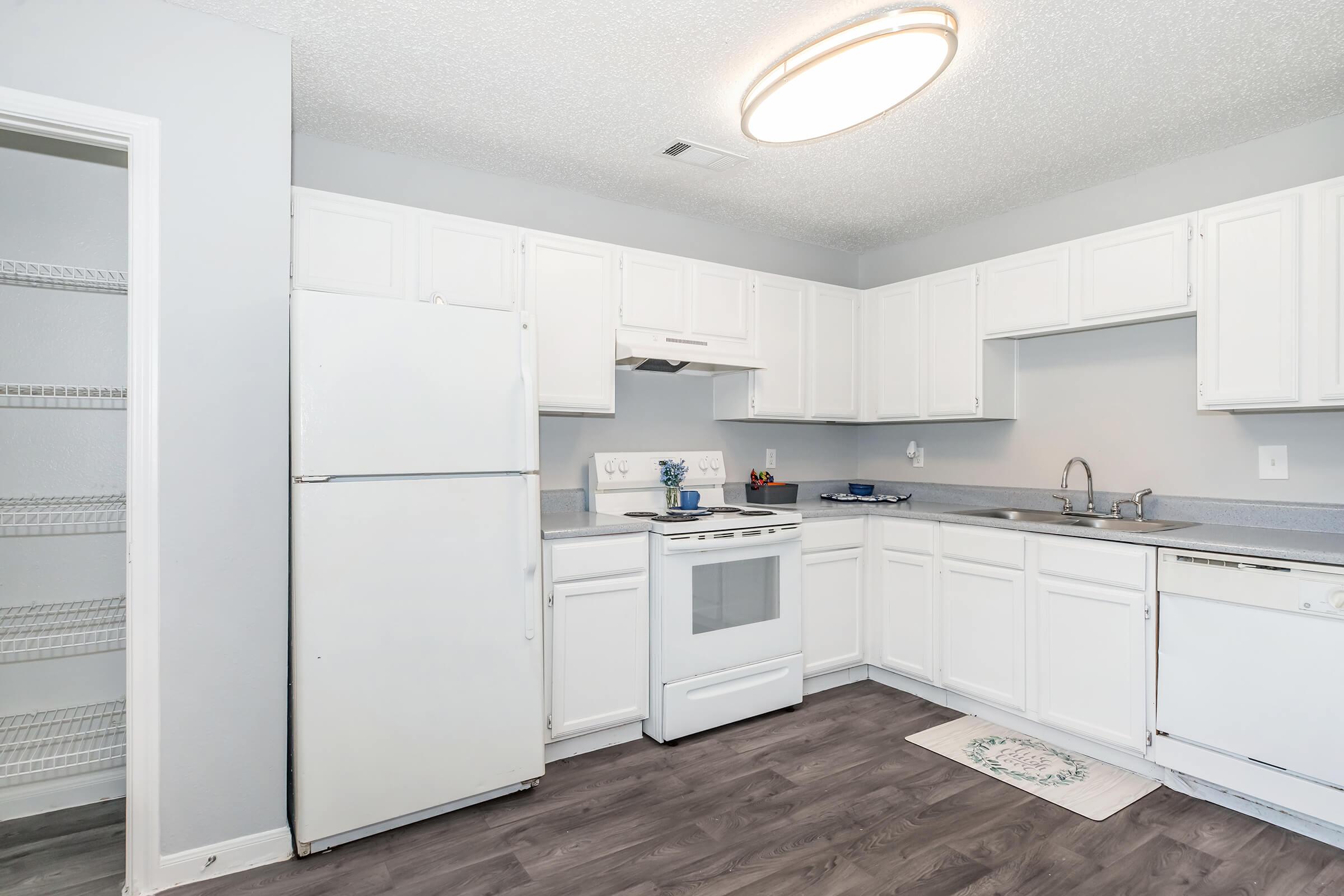 A clean, modern kitchen featuring white cabinetry, a white refrigerator, a stove, and a sink. The space has gray walls and a light-colored floor. A pantry shelf is visible in the corner, and a small rug lies in front of the sink. A circular ceiling light provides overhead illumination.