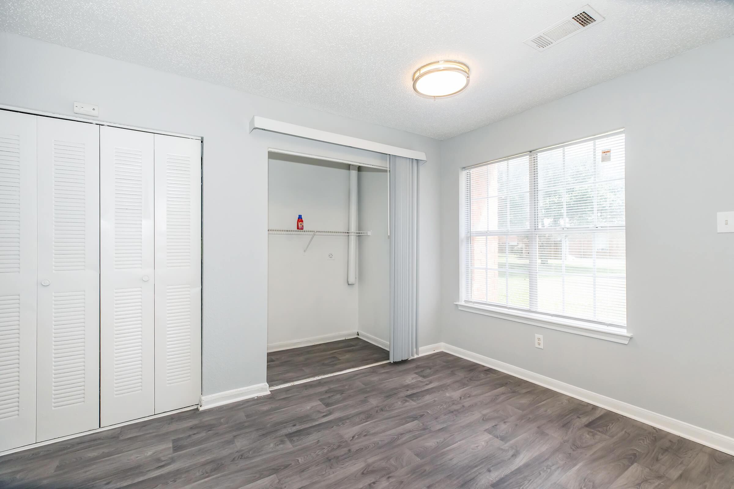 A well-lit room with gray walls and a light-colored laminate floor. It features a large window with a view of greenery outside, a closet with sliding doors, and a simple ceiling light. The space is uncluttered and ready for furnishings, creating a clean and inviting atmosphere.