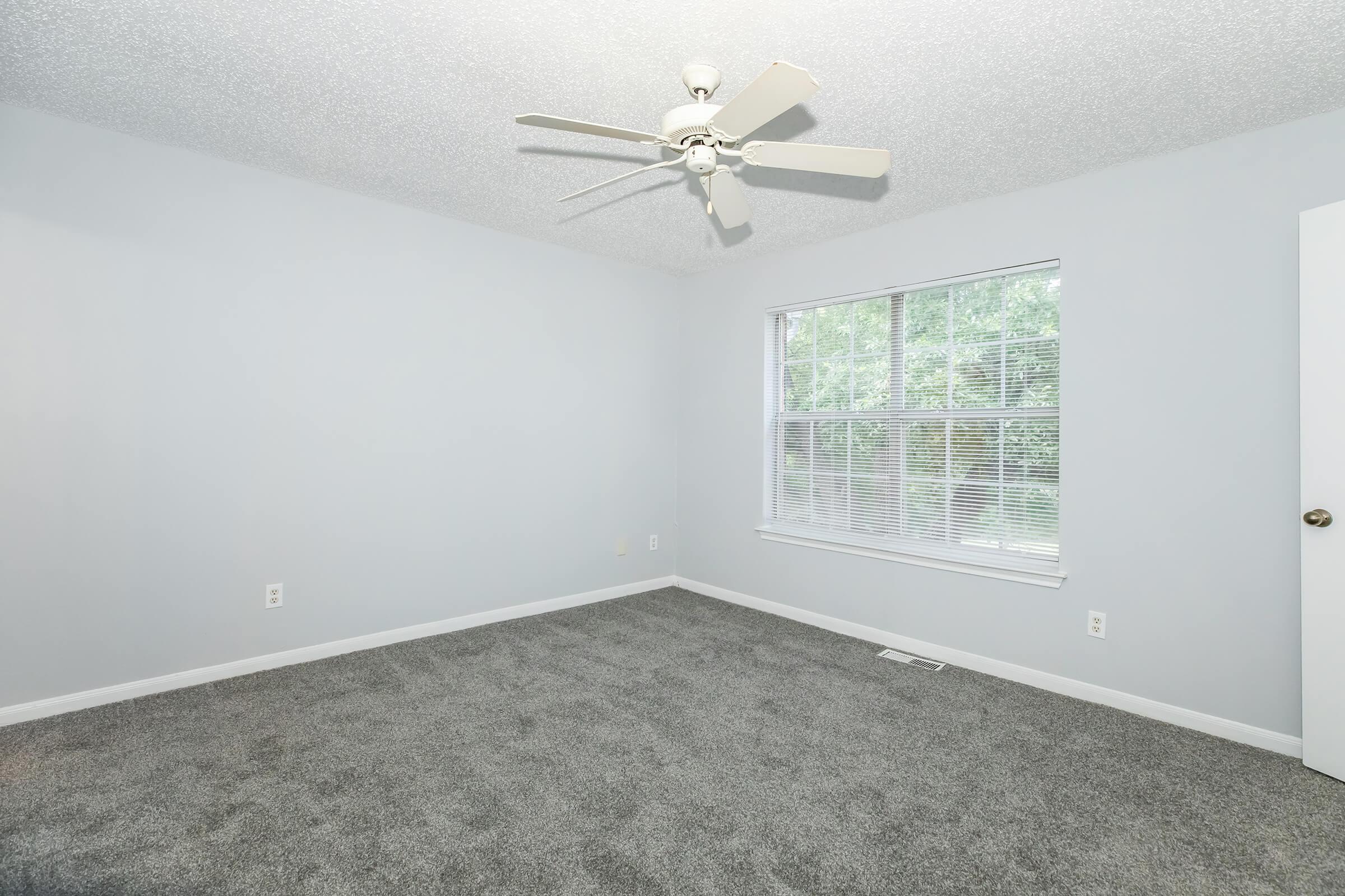 A minimalistic room with light gray walls and a ceiling fan. The floor is covered with soft gray carpet. A large window with blinds allows natural light to enter, and a white door is visible on the right side, contributing to the bright, airy feel of the space.