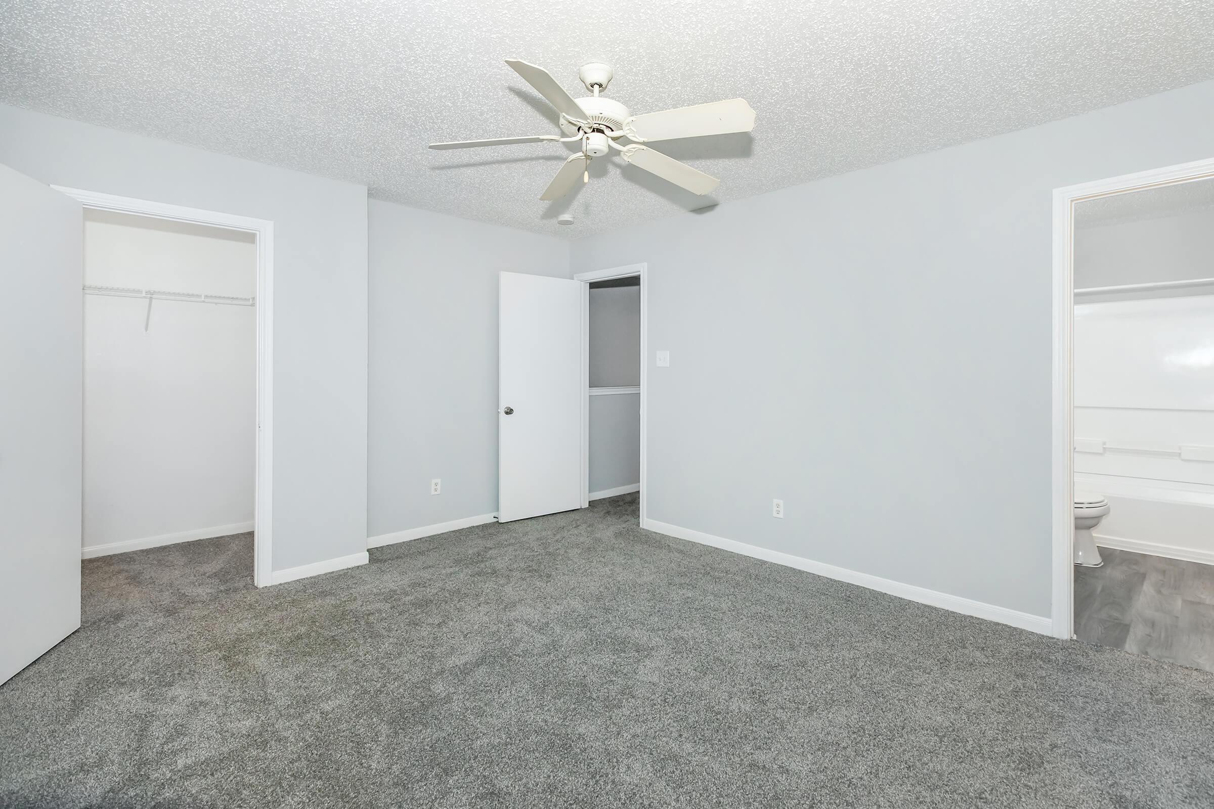 A well-lit, empty room featuring light gray walls and plush, gray carpeting. A ceiling fan is mounted overhead. There are two doors on the right side, one leading to a bathroom and the other to a hallway. A closet is visible on the left wall, and the overall space is spacious and clean.