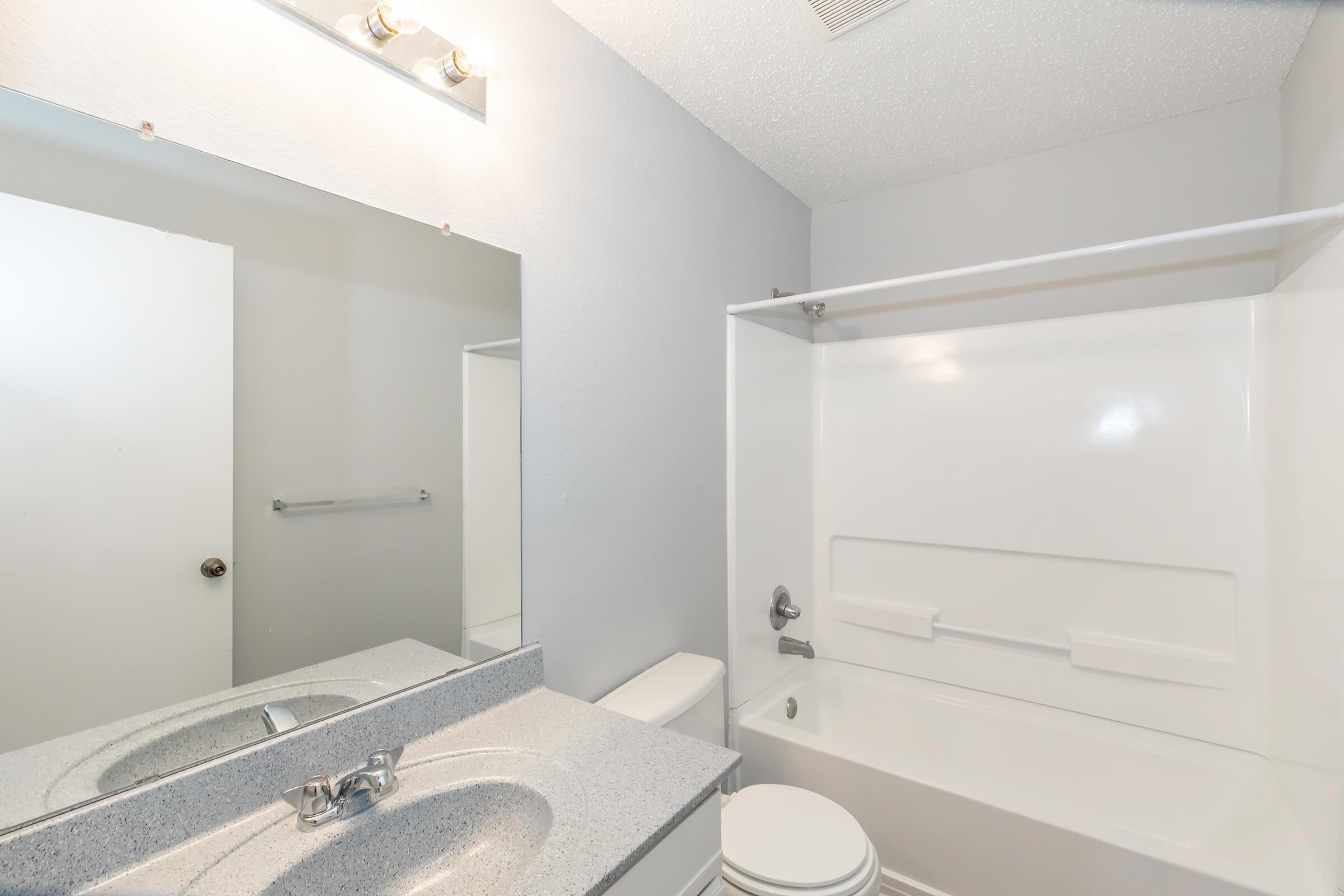 A clean and modern bathroom featuring a white bathtub with a shower curtain, a large mirror above a gray countertop sink, and a white toilet. The walls are painted in light gray, providing a bright and spacious feel. A towel rack is visible next to the mirror.