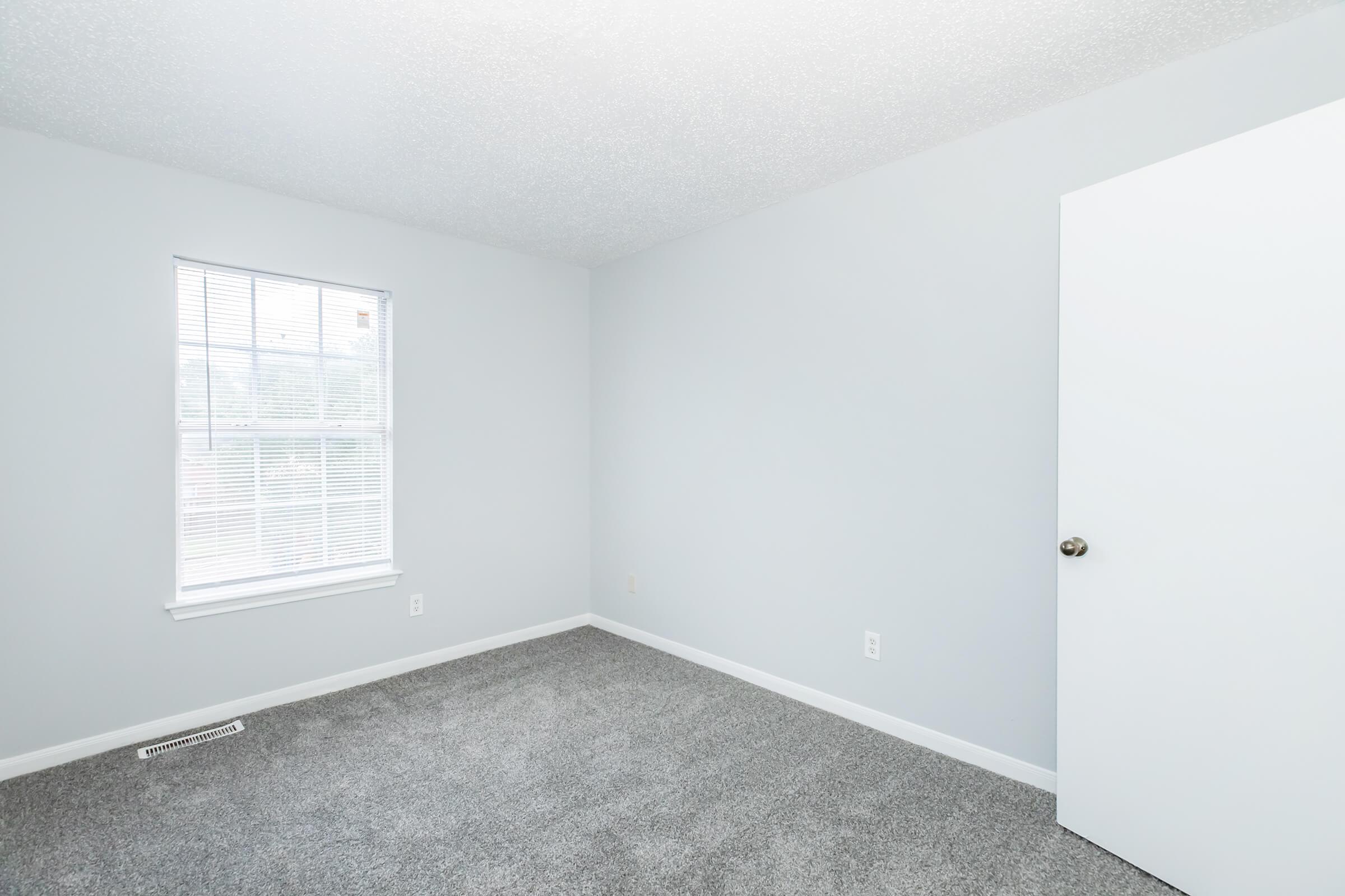 Empty room with light gray walls and a light gray carpet. A window with white blinds allows natural light in, and there is a plain white door on the right side of the image. The room has minimal furnishings, creating a clean and open space.