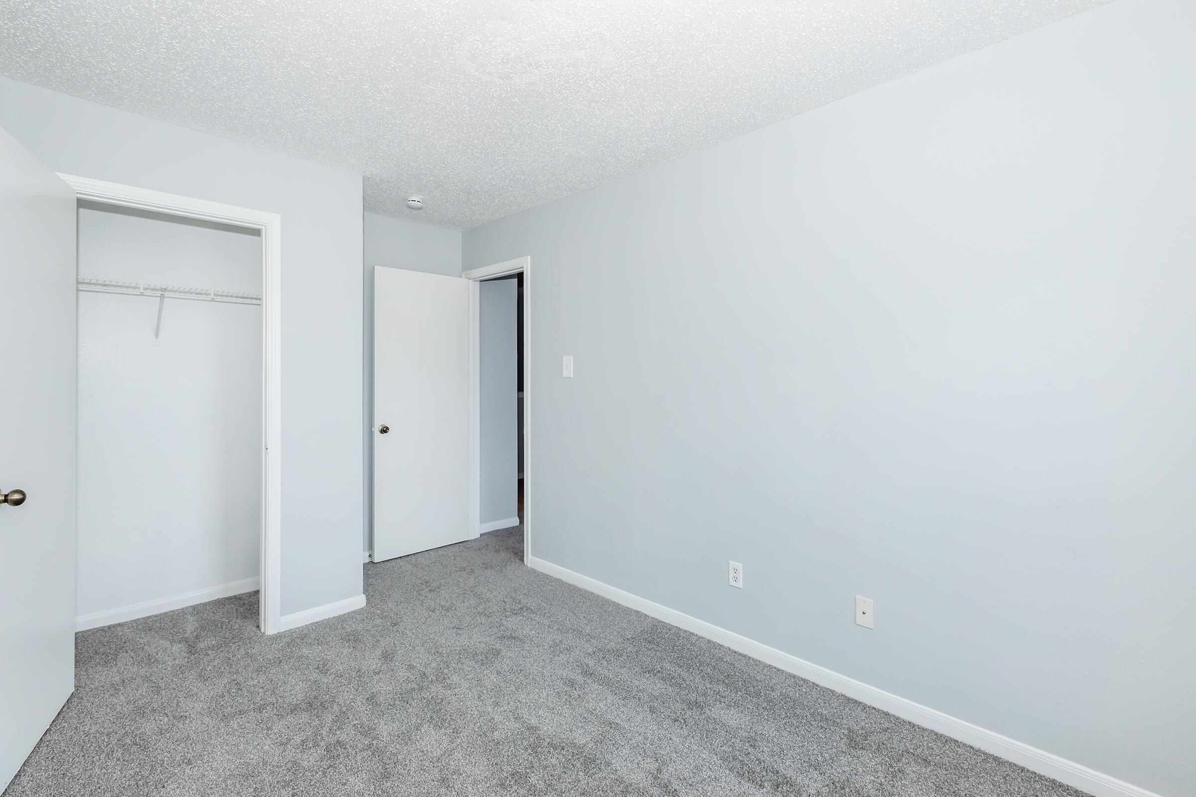 A well-lit, empty room with light gray walls and carpet. There are two white doors: one leading to a closet and another to an adjacent space. The overall decor is simple and modern, with no furniture present. Natural light is coming in from an unseen window, creating a spacious feel.