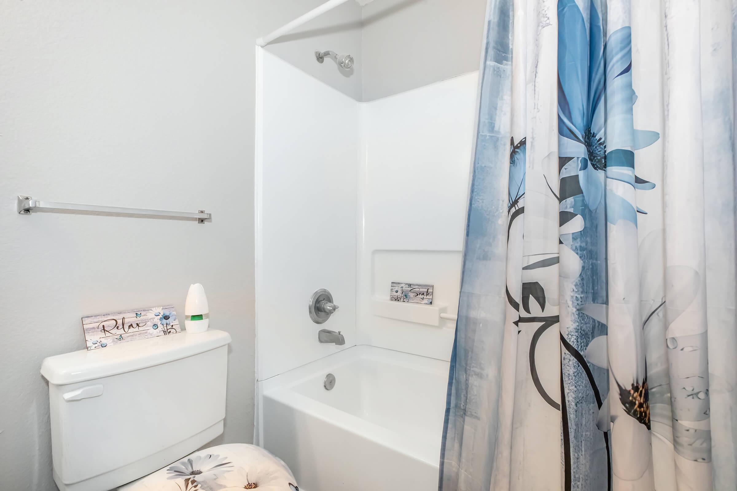 A small bathroom featuring a white bathtub with a shower curtain adorned with blue flowers. The toilet is next to the bathtub, and there's a towel rack on the wall. A decorative item with the word "Relax" is visible on a shelf, adding a touch of calmness to the space.