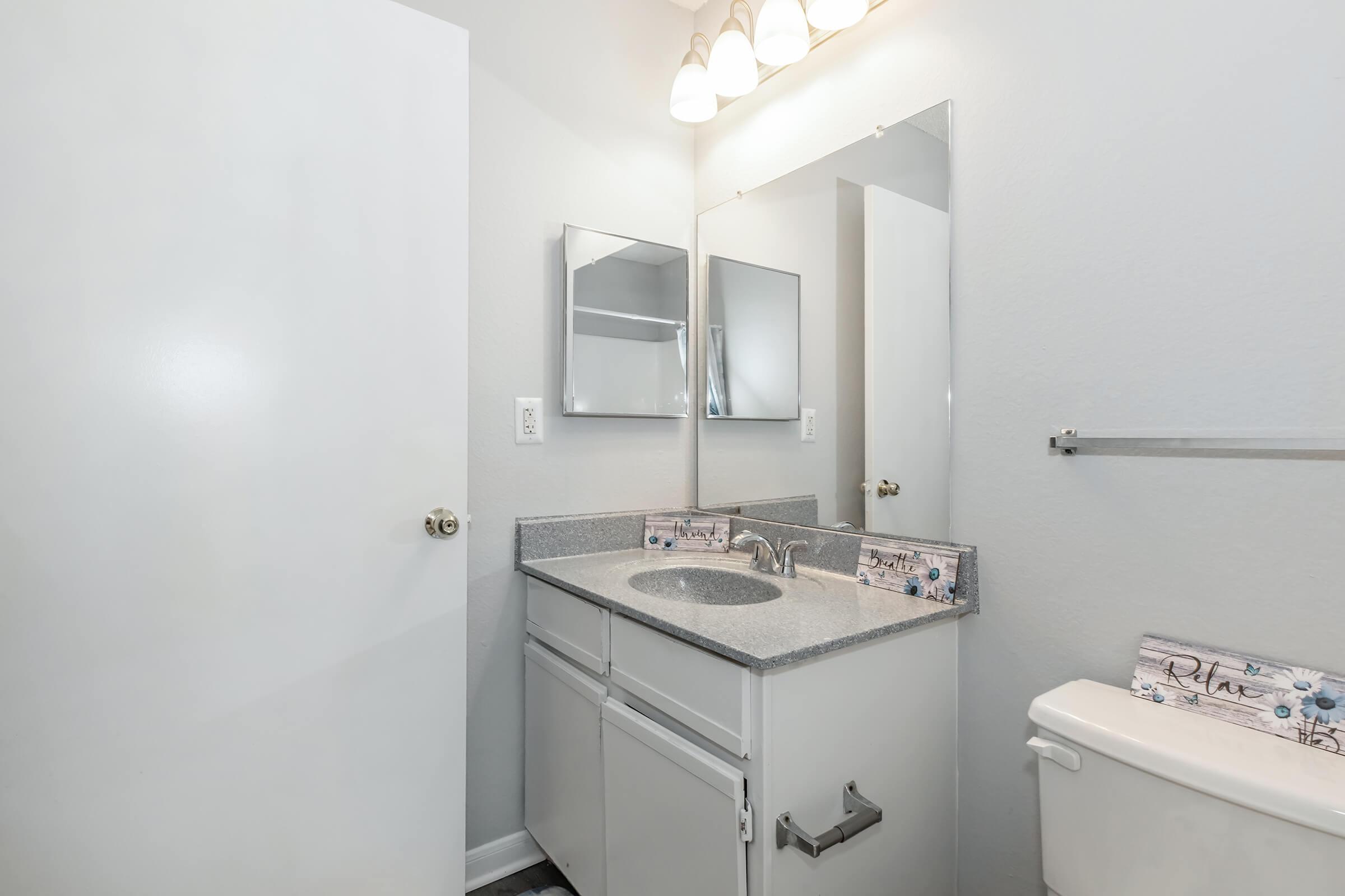 A small bathroom featuring a white cabinet with a granite countertop and sink, two mirrors above the sink, a toilet on the right, and a towel bar on the wall. The walls are painted light gray, and the lighting fixtures consist of three bulbs above the mirrors.