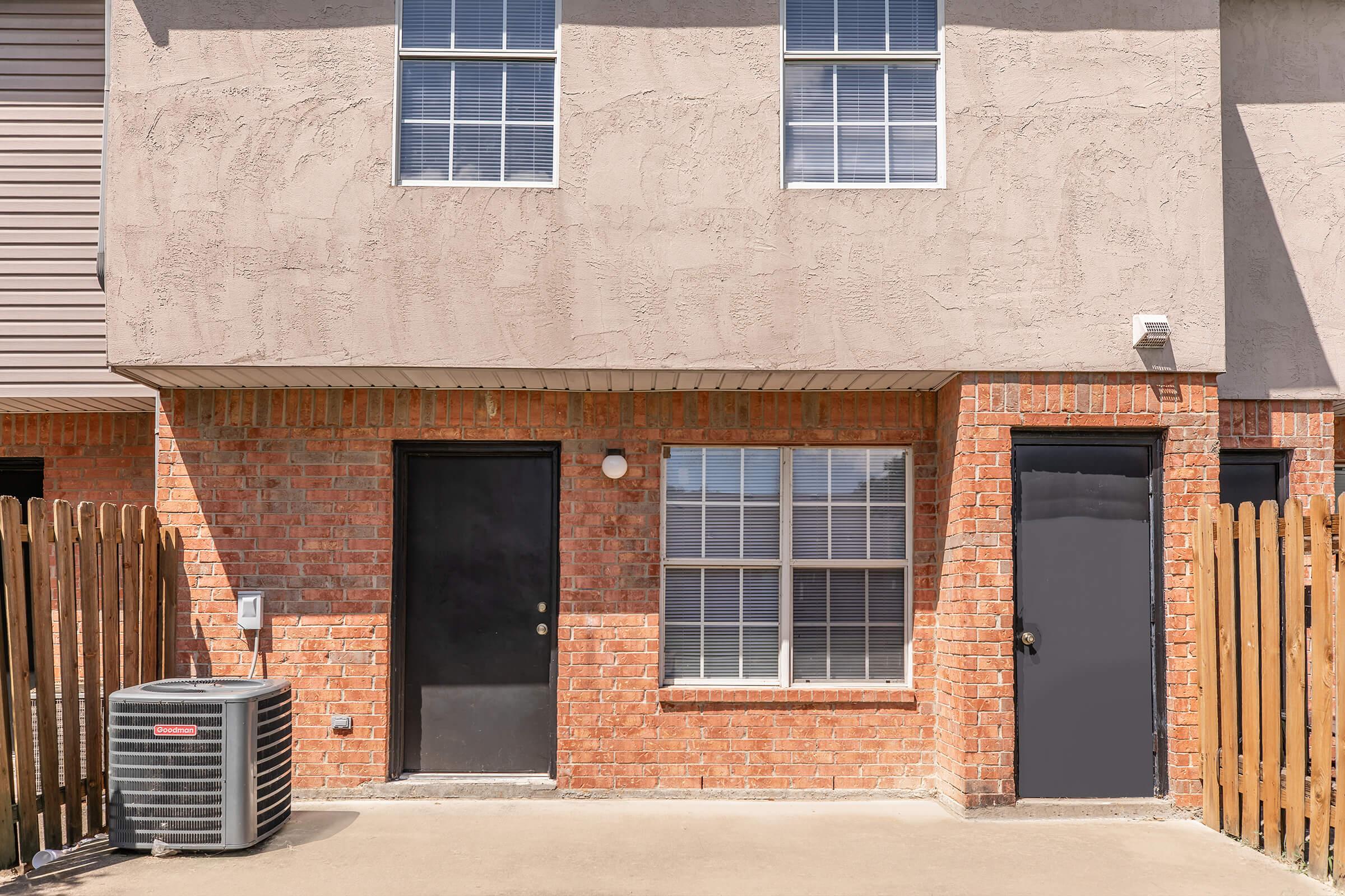 Exterior view of a residential building featuring a brick facade with two black doors and a large window. An air conditioning unit is positioned to one side, and a wooden fence surrounds the entrance area. The building has a light-colored textured upper section and ample natural light coming through the window.