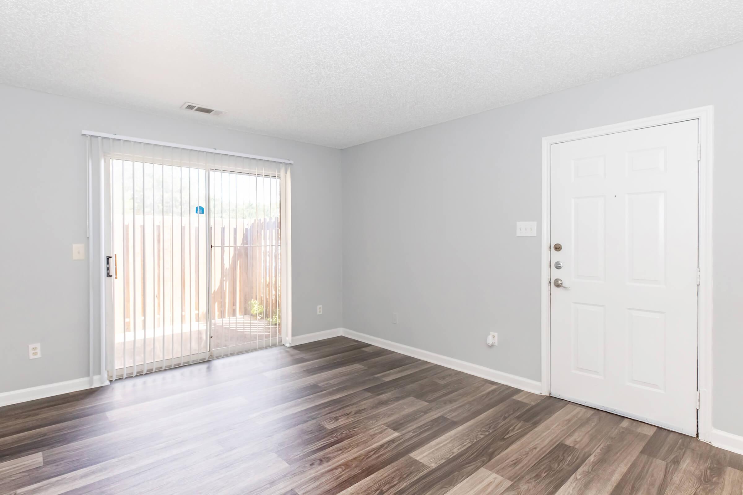 Spacious, empty room featuring light-colored walls and modern vinyl flooring. Double sliding glass doors lead to an outdoor area, creating a bright and airy atmosphere. A white door provides access to another space, offering potential for various uses.