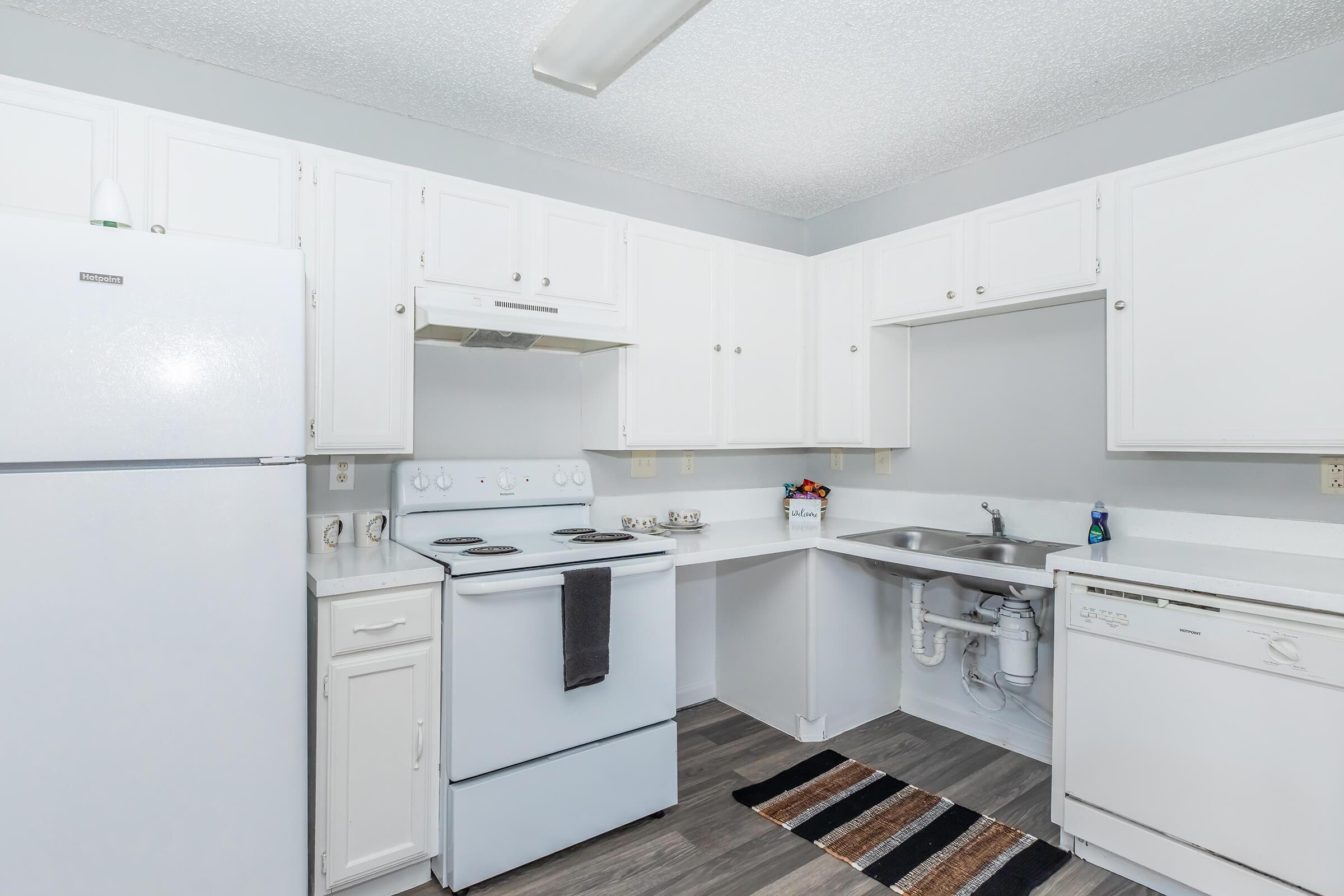 A clean and modern kitchen featuring white cabinets, a white refrigerator, and a white stove with an oven. The countertop is light-colored with a sink and kitchen mat. Gray walls and wooden flooring complete the look, creating a bright and functional space.