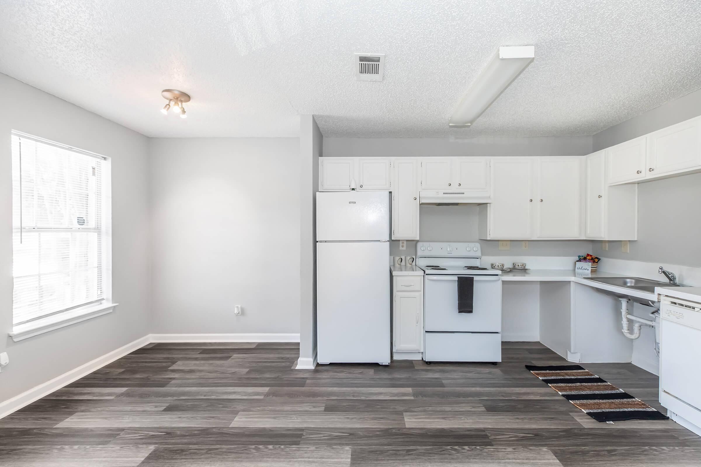 A bright and modern kitchen featuring white cabinetry, a refrigerator, an oven, and a sink. The flooring is a dark wood laminate, and there’s a small rug in front of the sink. Windows allow natural light to fill the space, enhancing the clean and open atmosphere.