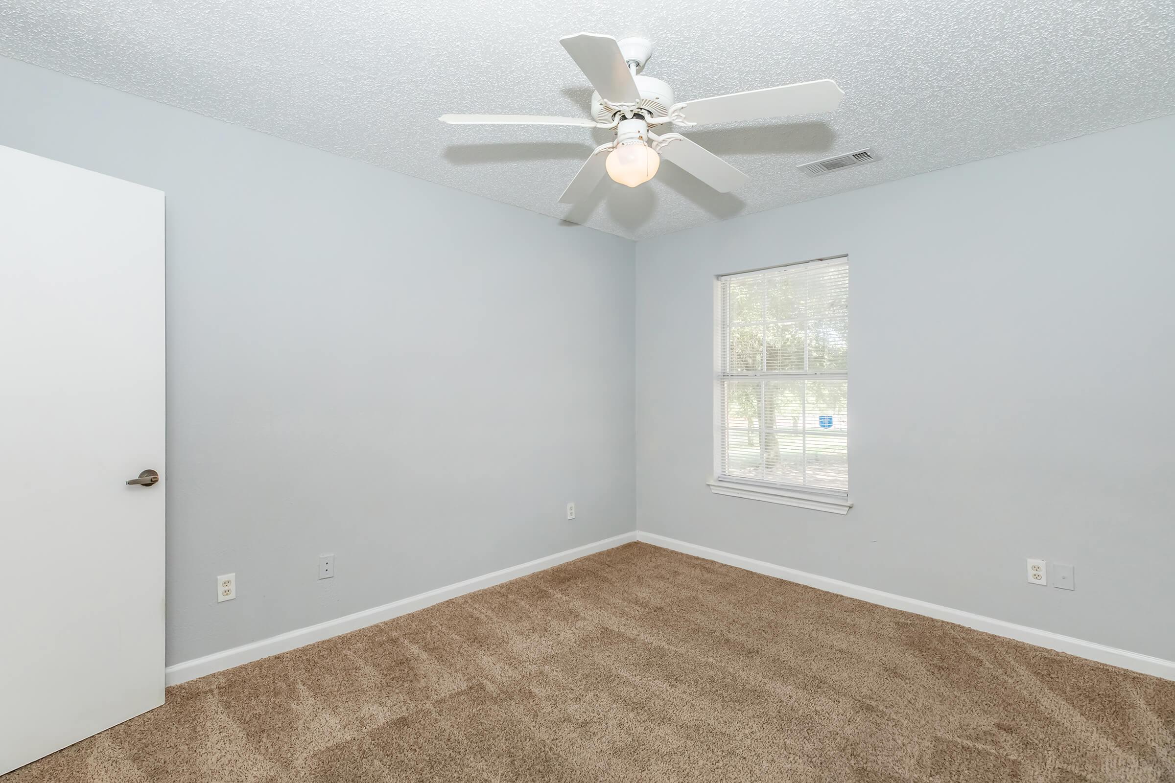 A light blue bedroom featuring a ceiling fan with a light fixture, a white door, a window with natural light, and beige carpet flooring. The walls are painted a soft blue, creating a calm atmosphere. The room is empty, highlighting its spacious and clean appearance.