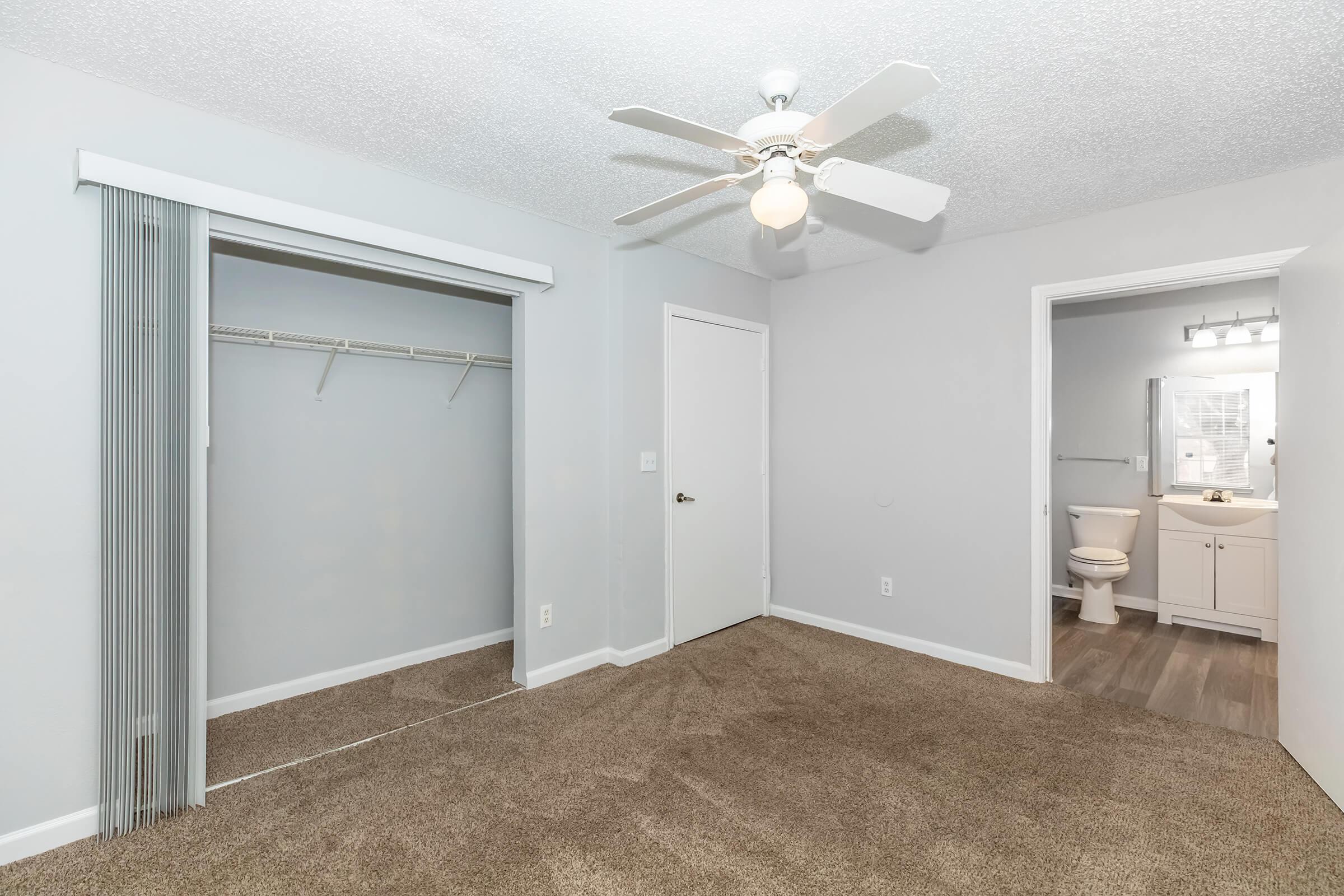 A spacious bedroom featuring light gray walls, carpet flooring, a ceiling fan, and a sliding closet. There is an adjoining bathroom visible through an open door, which includes a sink and toilet. Natural light enters the room through a window, creating a bright and inviting atmosphere.