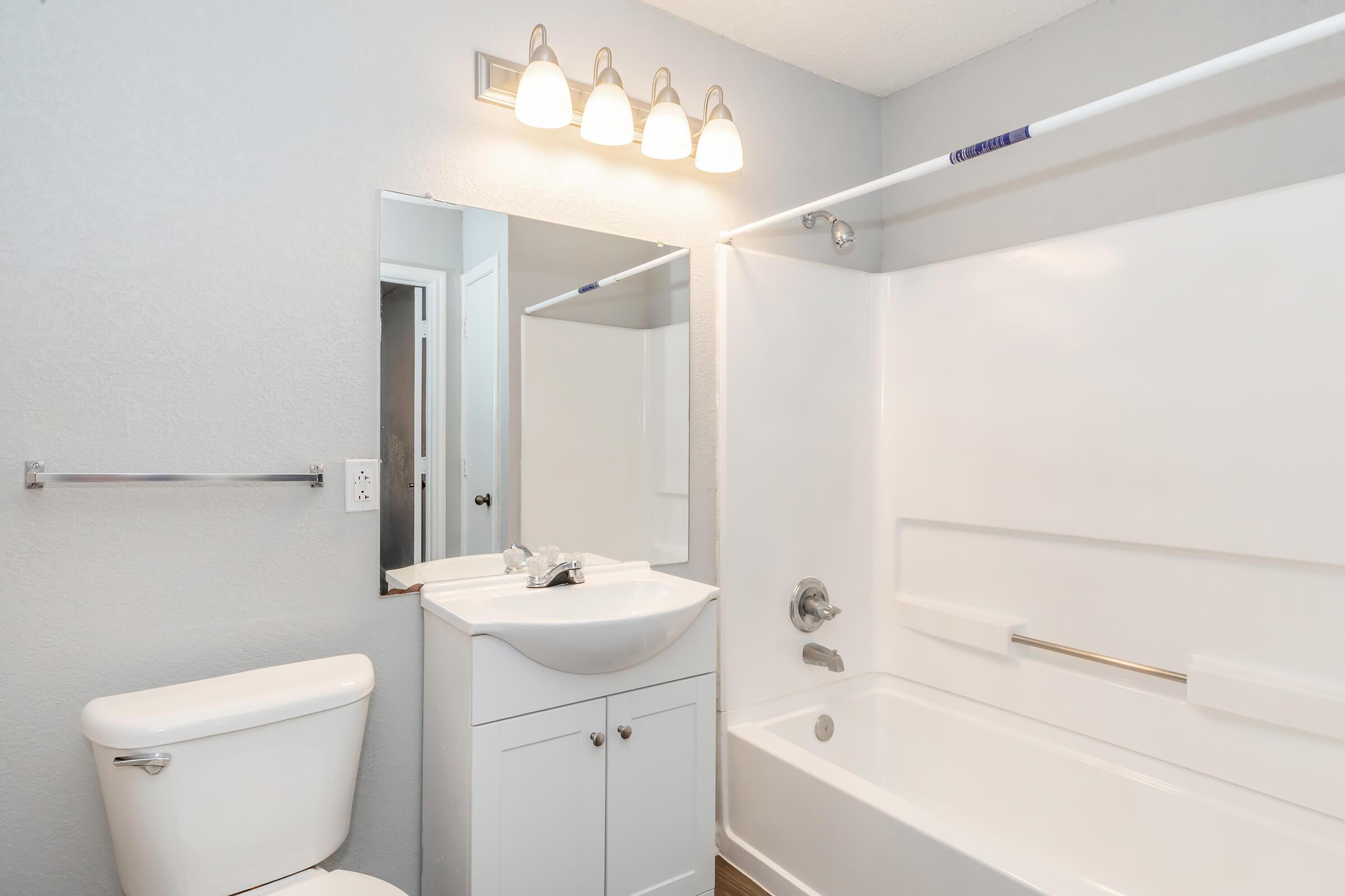 A clean, modern bathroom featuring a white toilet, a vanity with a sink and mirror, and a shower-tub combination. The walls are painted a light gray, and there are three light fixtures above the mirror. A towel bar is mounted beside the sink, adding to the functional design.