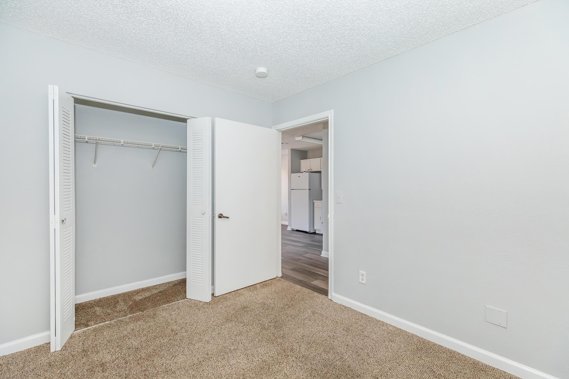 A vacant bedroom with light gray walls and a carpeted floor. A closet with sliding doors is on the left, and the room opens to a kitchen area in the background, visible through an open door. The space is well-lit and clean, suggesting a modern, simple design.