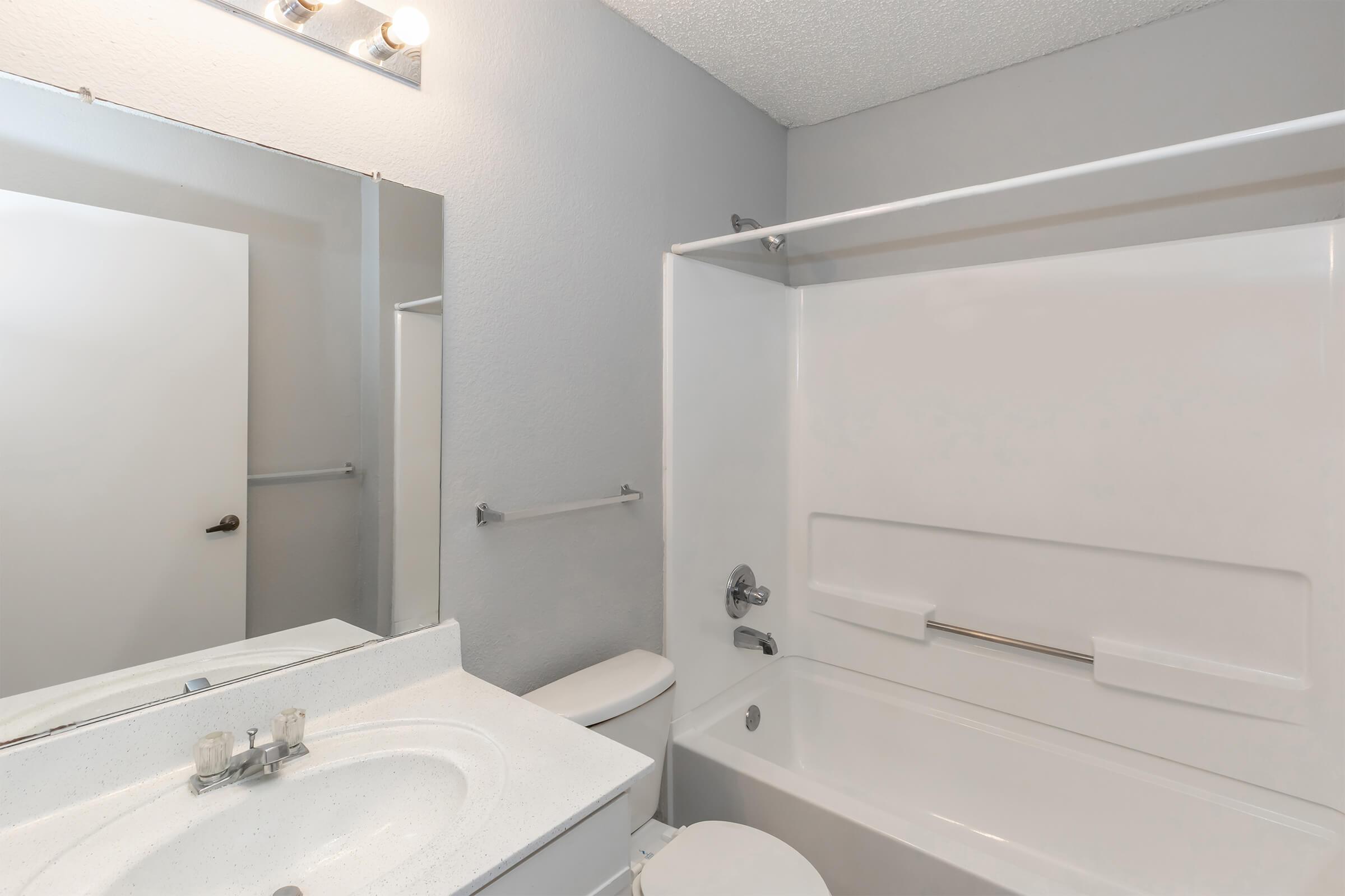 A clean and modern bathroom featuring a white bathtub and shower combination, a white sink with a countertop, a mirror above the sink, and a light fixture above the mirror. The walls are painted gray, and there is a white door visible in the background.