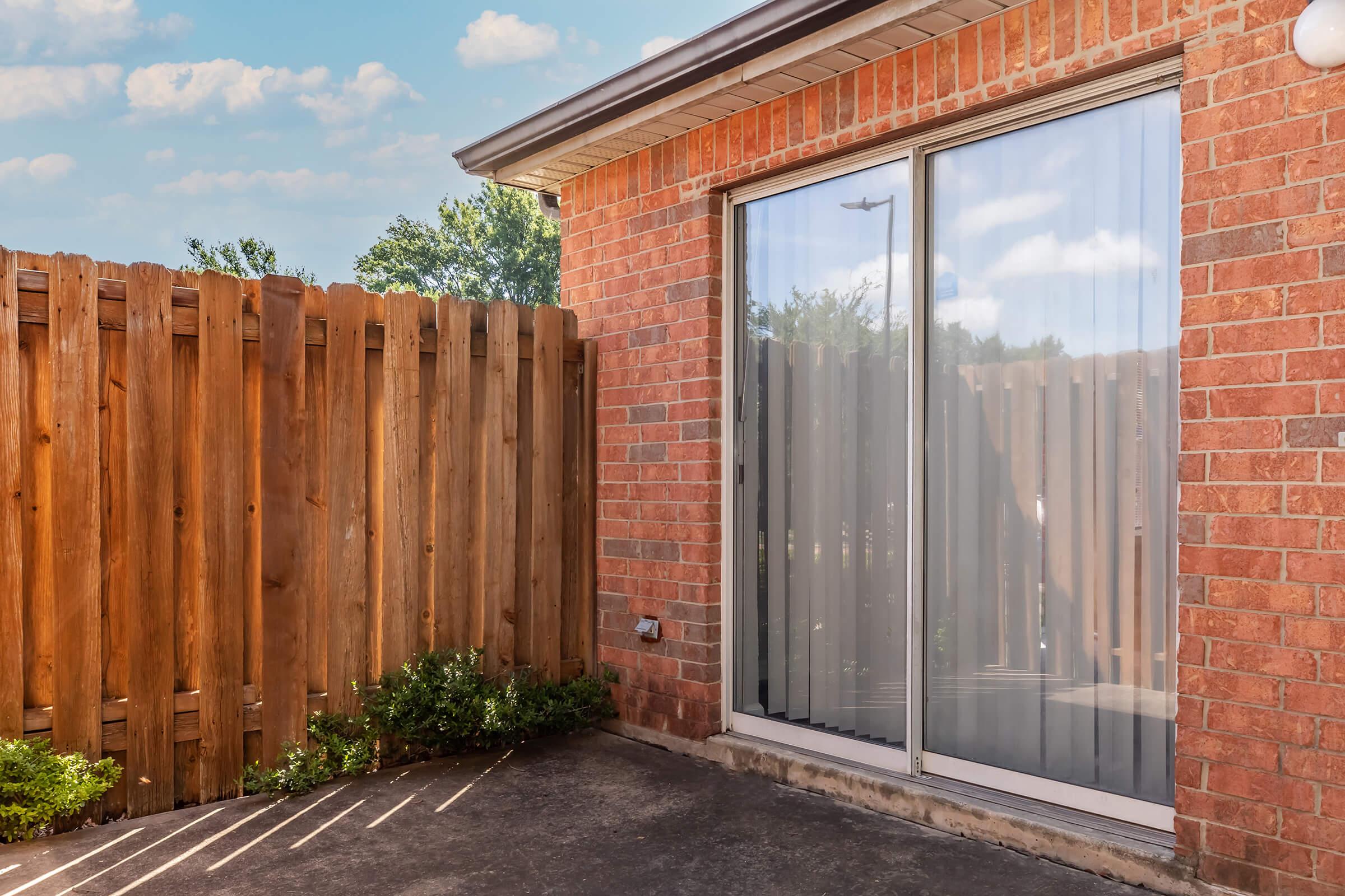 A brick wall with a sliding glass door leading to a patio area, surrounded by a wooden fence. There is greenery at the base of the fence, and the sky is blue with a few clouds visible in the background. Sunlight casts shadows on the concrete.
