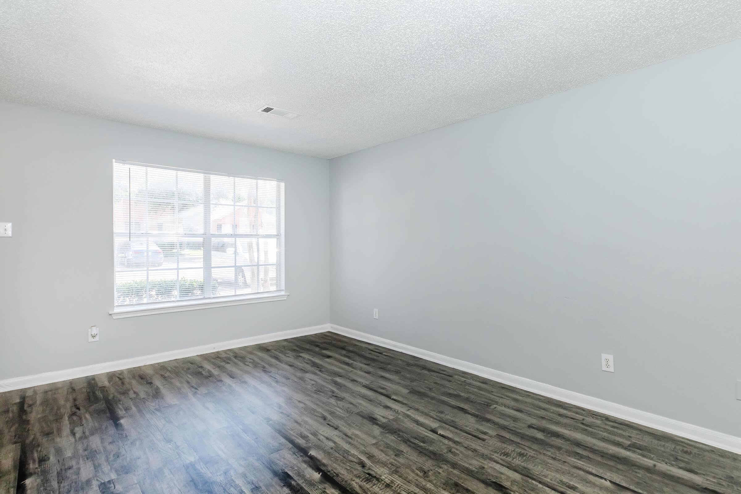 A spacious, empty room with light gray walls and a textured ceiling. The flooring is dark wood laminate, and there's a large window allowing natural light to fill the space, offering a view of the outdoors. The room has no furniture, creating a bright and open atmosphere.