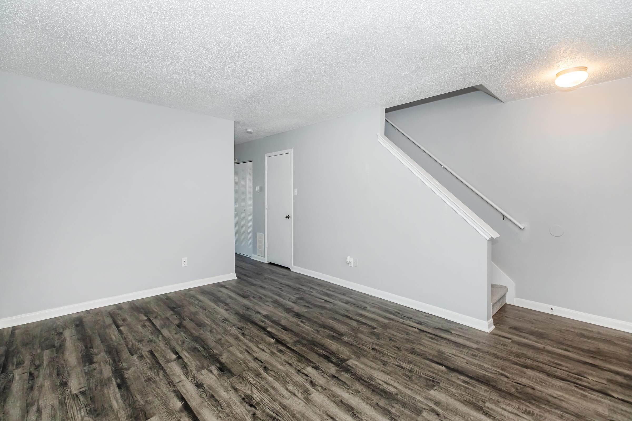 A spacious, modern living area with light gray walls and dark wood-like flooring. The room features a staircase leading to an upper level, a ceiling light fixture, and an open layout with access to a hallway. Natural lighting brightens the space, creating a welcoming atmosphere.