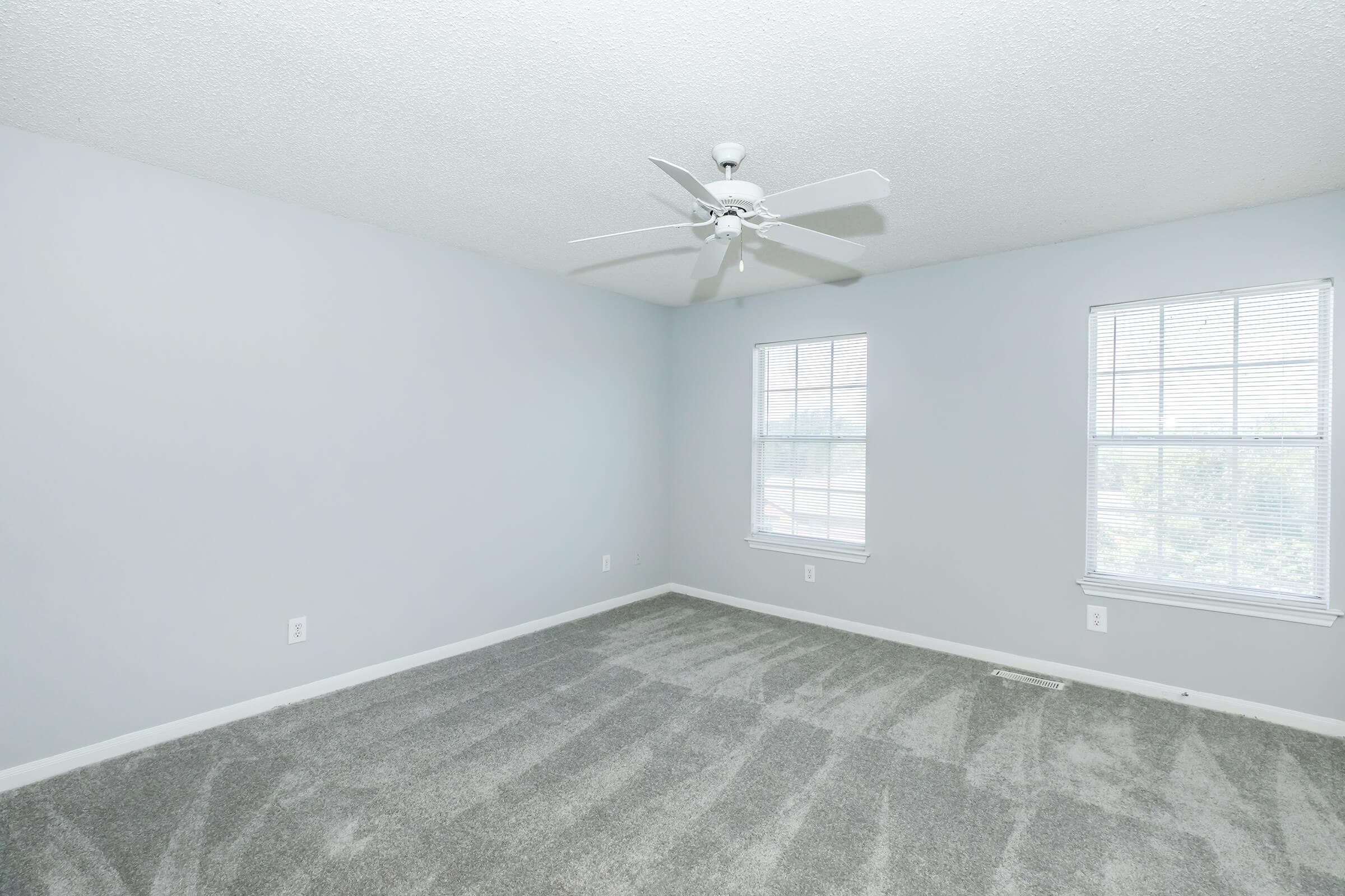 Spacious and empty room with light gray walls and carpet flooring. Two windows allow natural light to fill the space, and a ceiling fan is mounted above. The overall atmosphere is bright and airy, ideal for personalization and decoration.