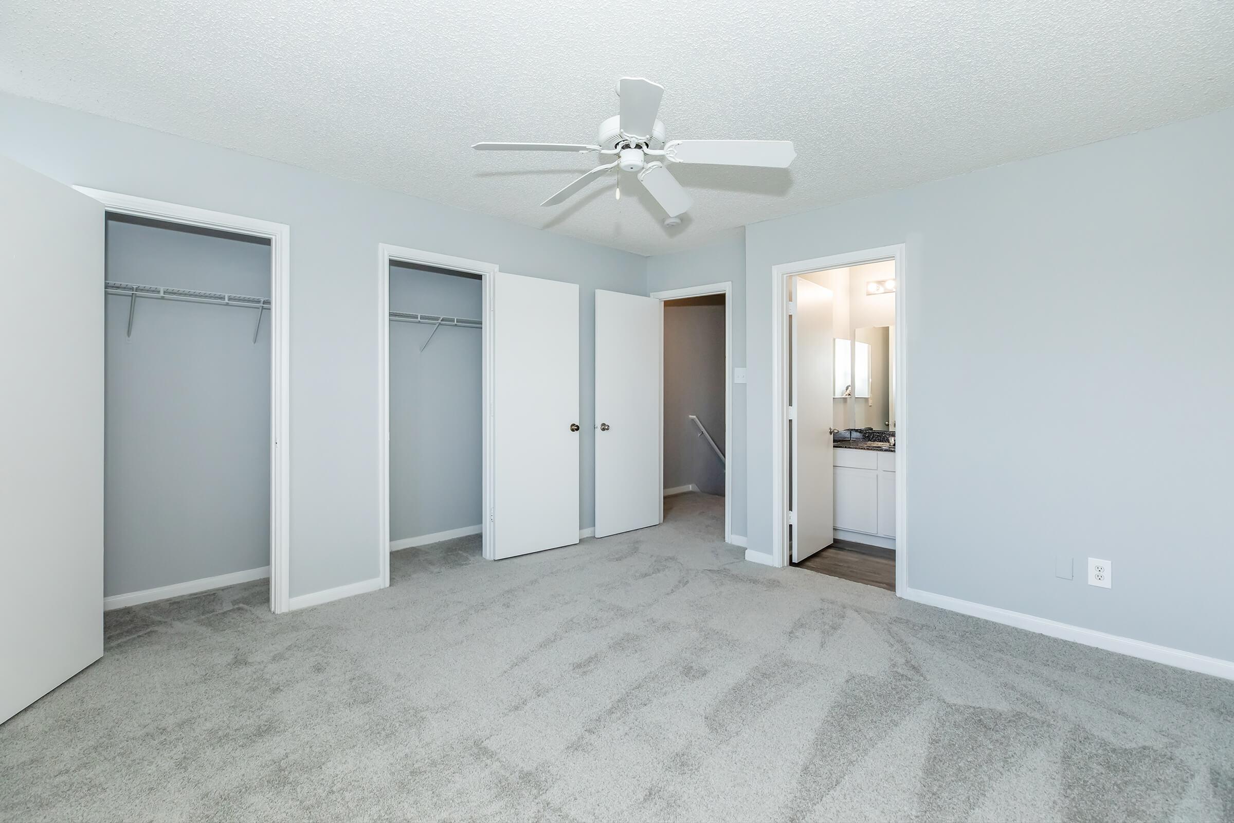 A spacious, light gray bedroom featuring carpeted flooring, a ceiling fan, and multiple doors leading to closets and a bathroom. The walls are painted a soft gray, creating a serene atmosphere. Natural light enters through the doors, enhancing the open feel of the room.
