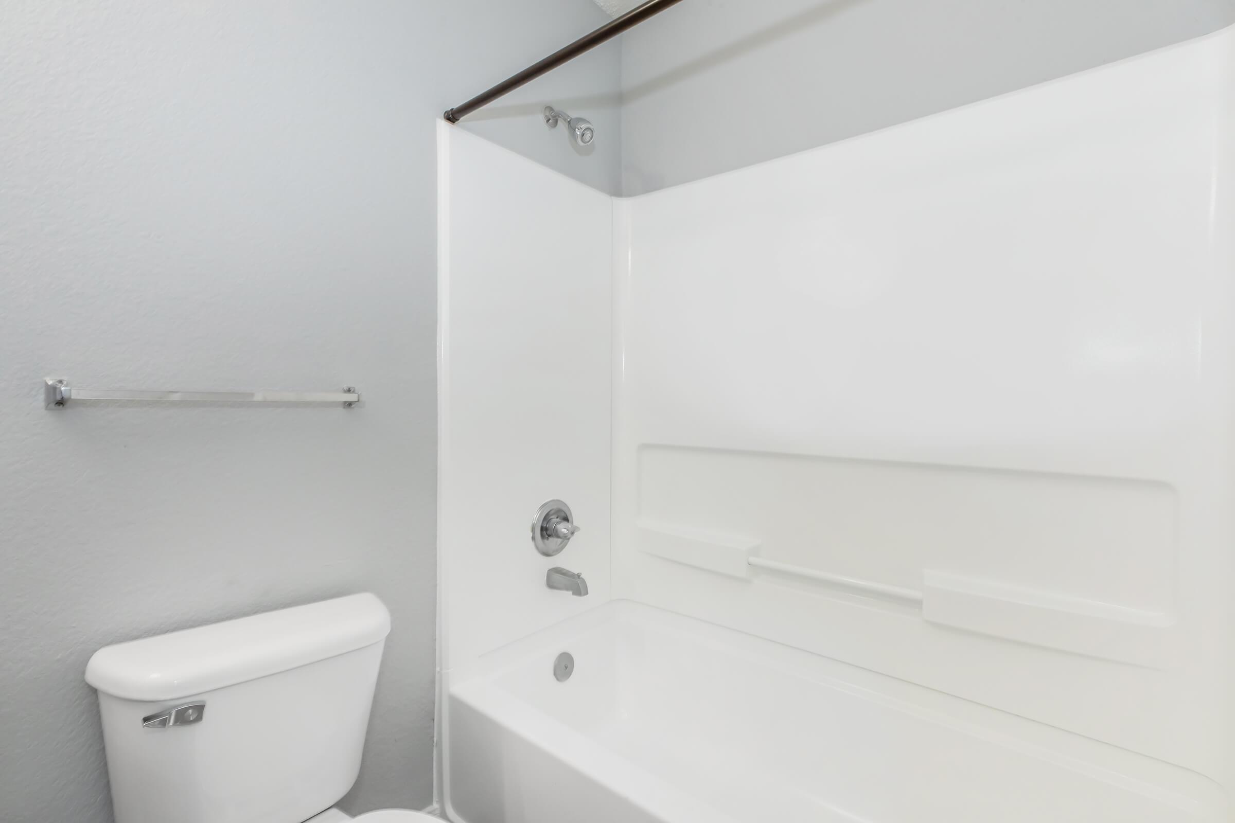 A clean, minimalist bathroom featuring a white bathtub with a glass shower door, a modern toilet, and a towel rack on a gray wall. The space is bright and uncluttered, emphasizing a simple and functional design.