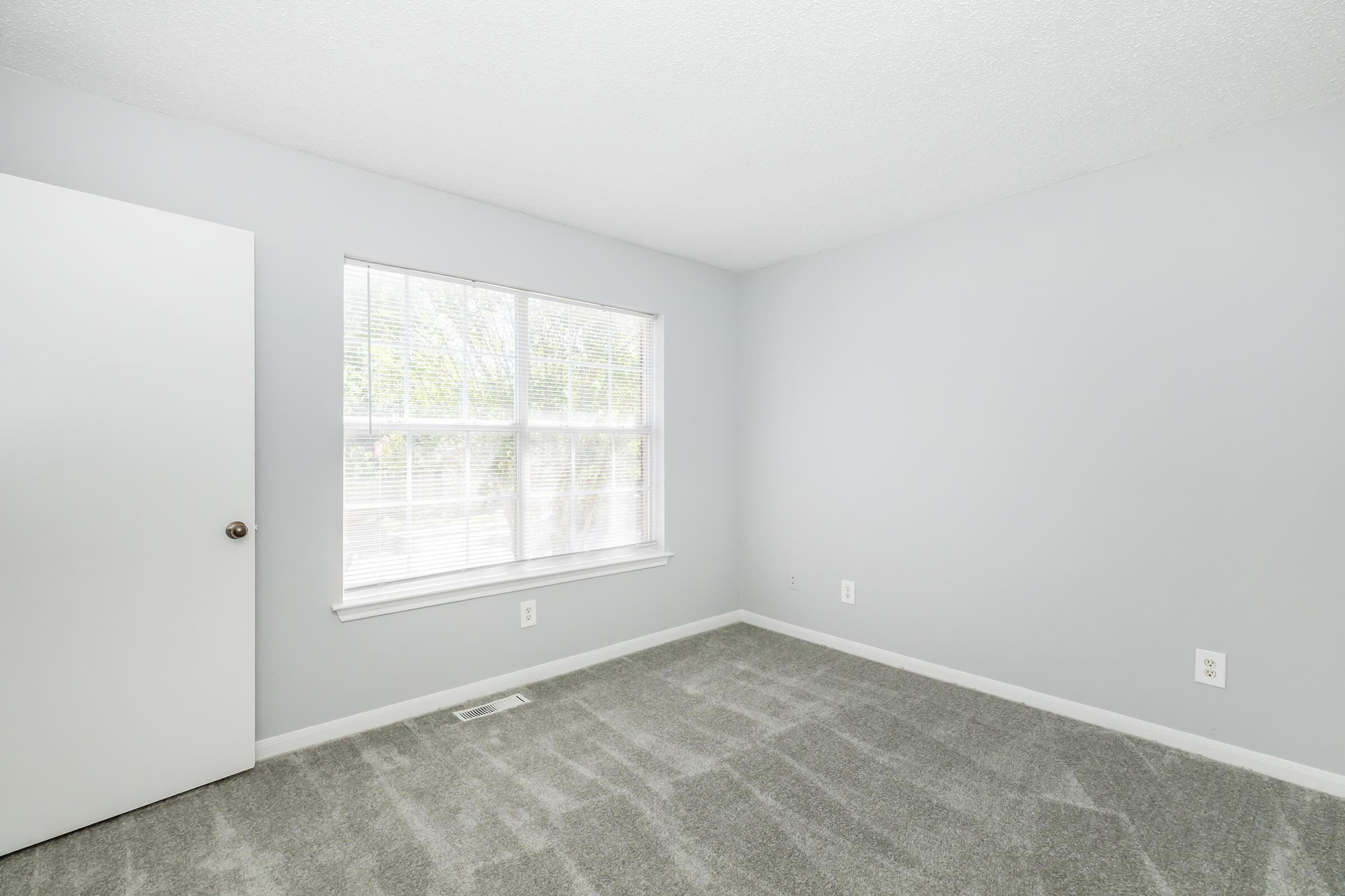 Empty room with light gray walls and gray carpet. A window with blinds allows natural light to enter, and there is a closed white door on the left side of the image. The room appears clean and unfurnished, providing a neutral space for potential use or decoration.