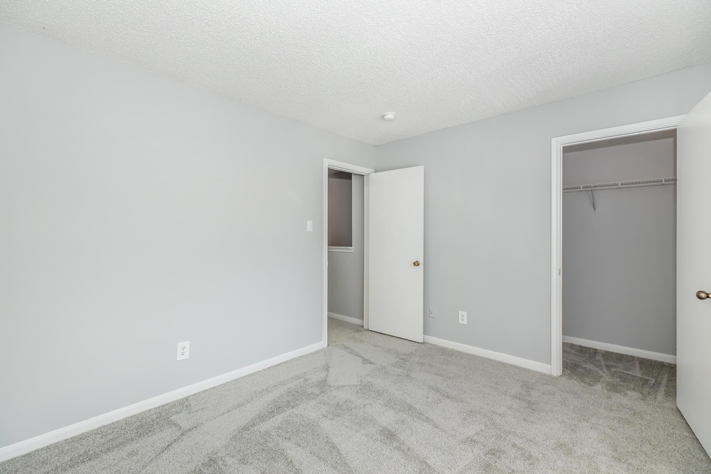 A bright, empty room featuring light gray walls and soft gray carpeting. Two doors are visible: one leading to a closet and the other to an adjacent space. Natural light comes from a small window, creating an open and spacious feel in the area.