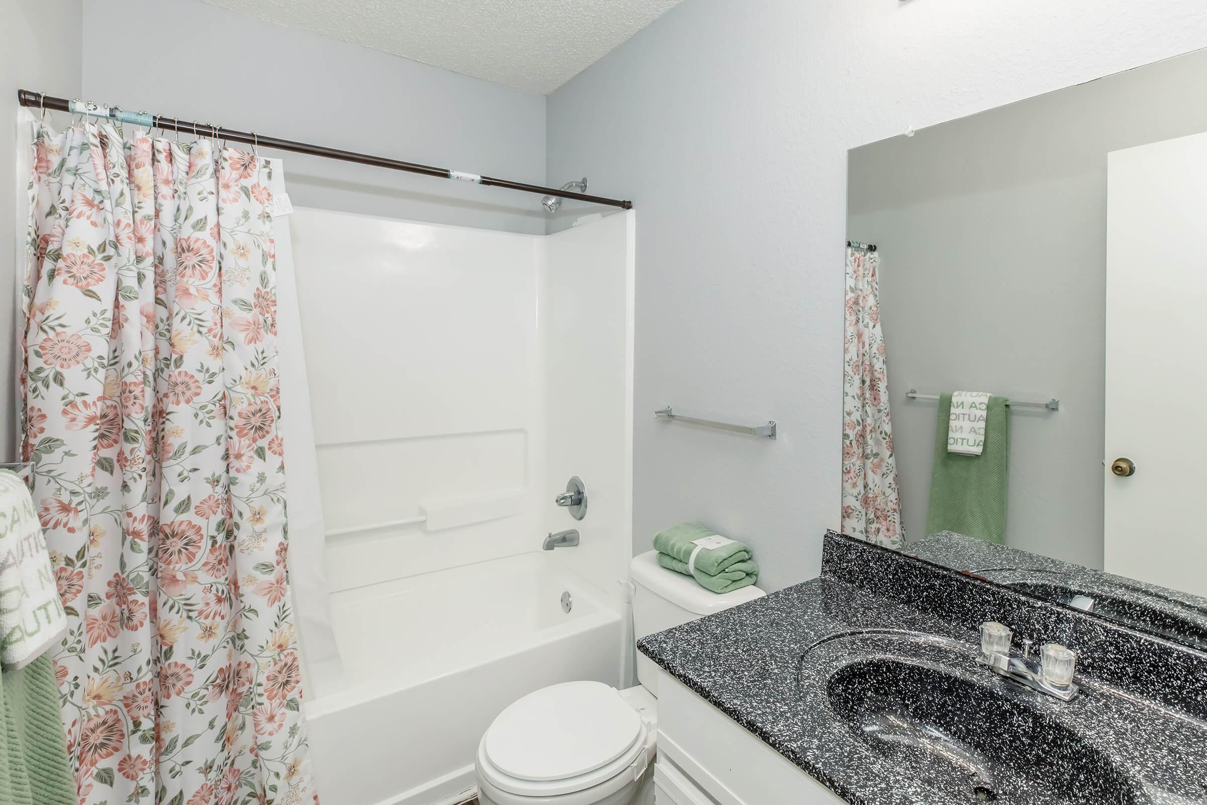 A clean, modern bathroom featuring a bathtub with a floral shower curtain, a dark granite countertop with a sink and mirror, a white toilet, and neatly folded towels. The walls are a light color, enhancing the space's brightness.