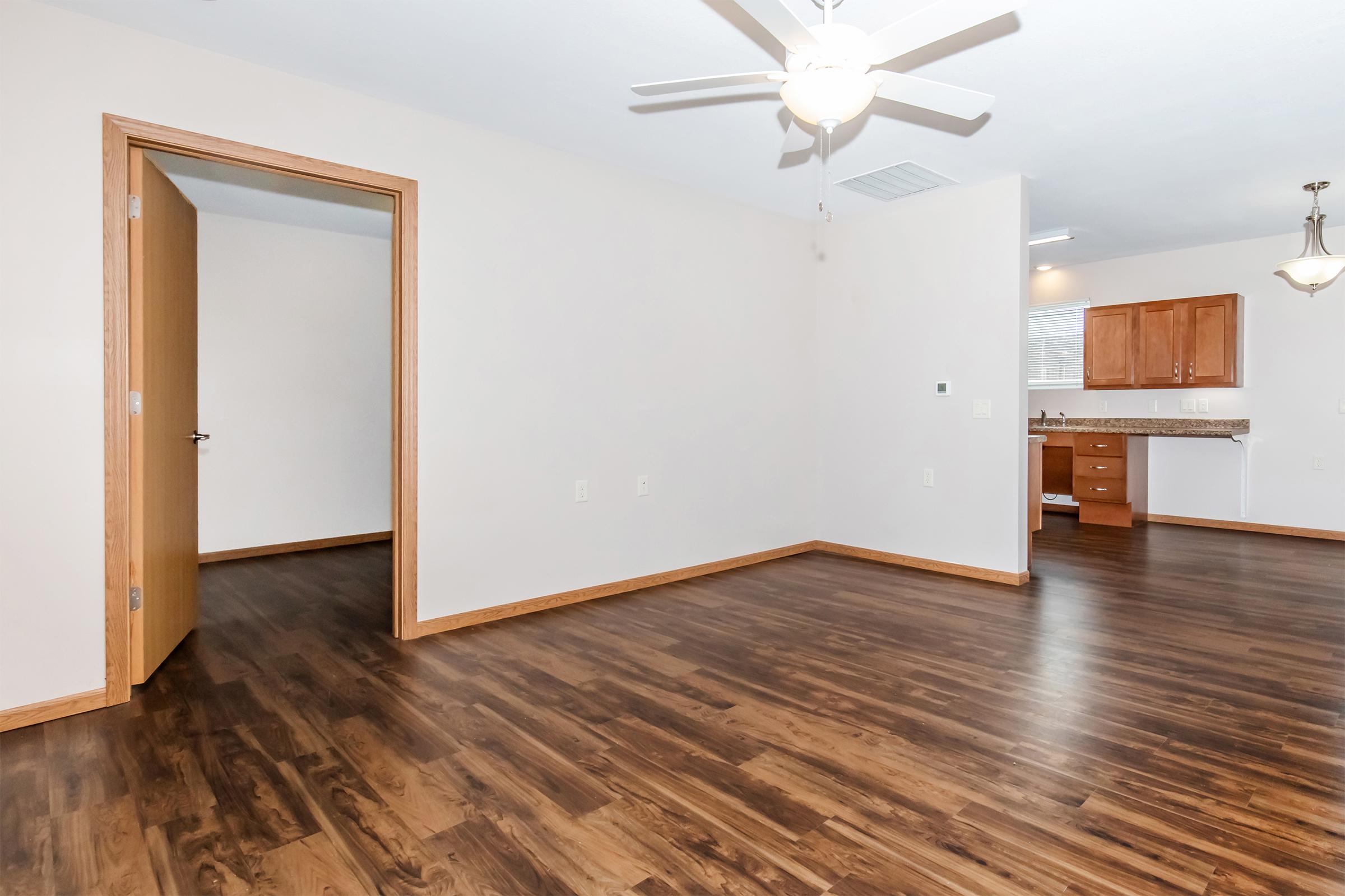 A spacious, well-lit living area featuring brown laminate flooring, a ceiling fan, and an open layout. To the left, there's a closed door leading to another room. In the background, a kitchen with wooden cabinets is visible. The walls are painted light, creating a bright atmosphere.