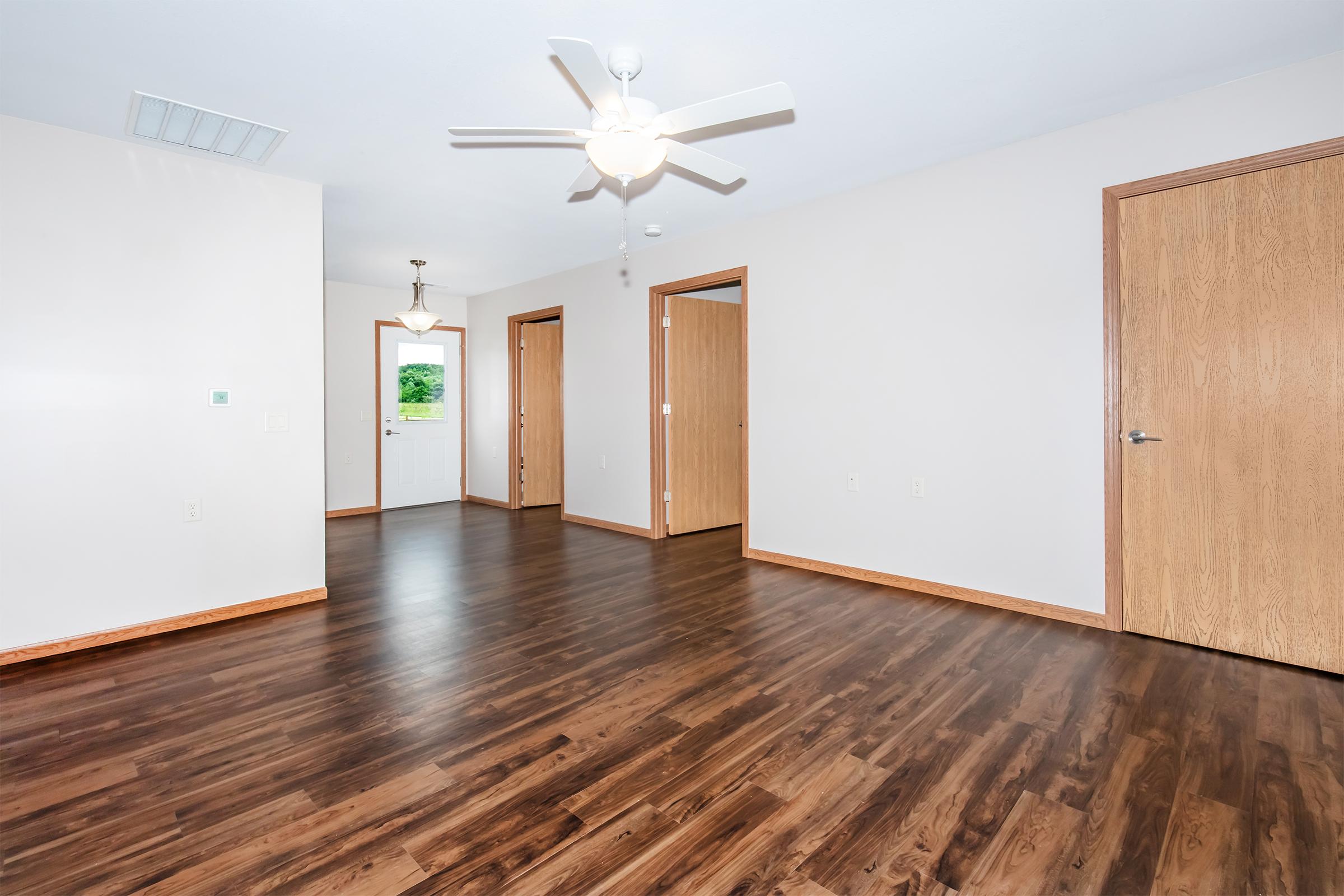 A spacious interior room featuring wooden floors, a ceiling fan, and light-colored walls. There are three doorways leading to other areas, with natural light coming from a window near the entrance. The room has a clean, modern aesthetic, suitable for various uses such as a living space or office.