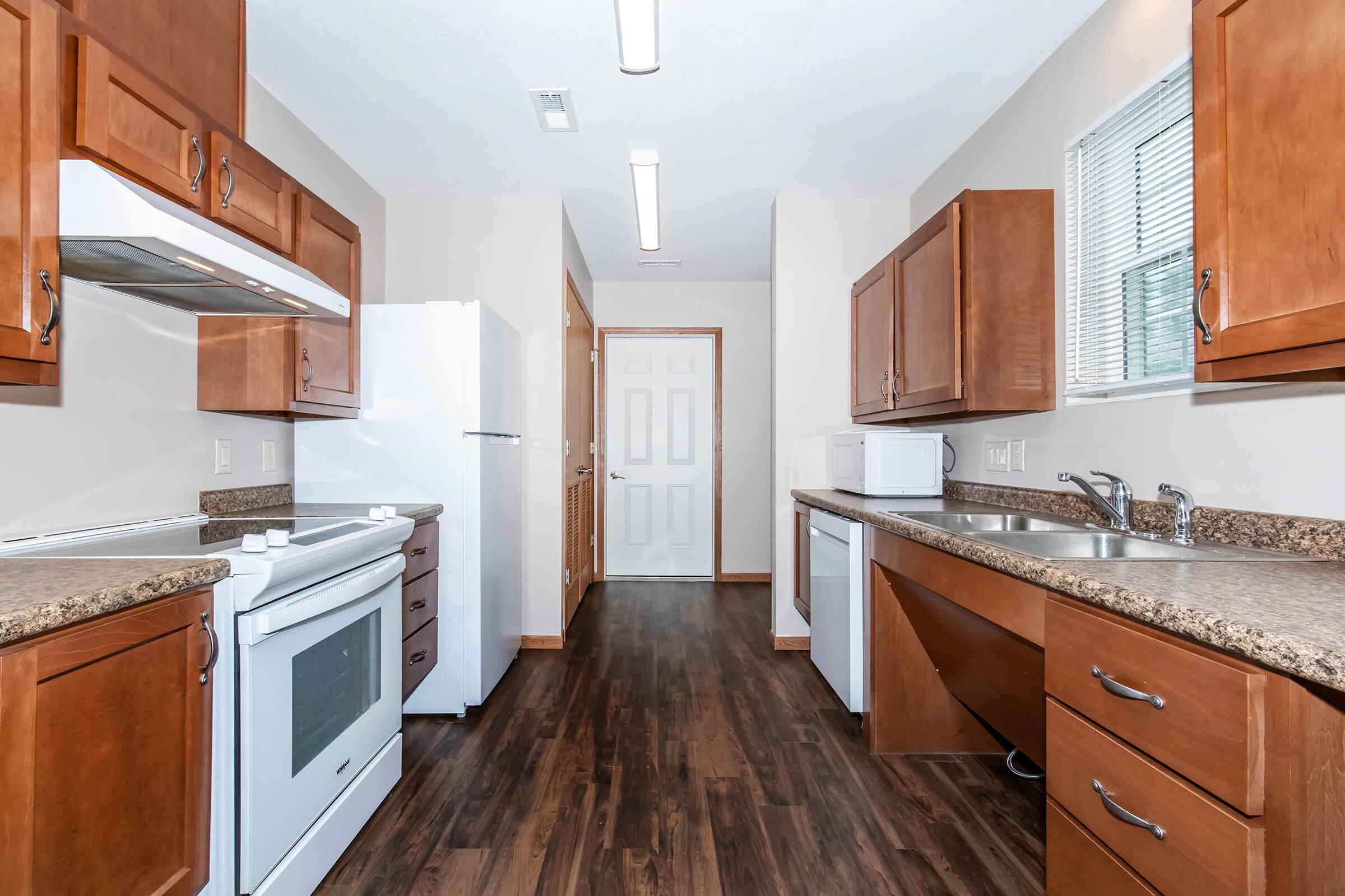 Modern kitchen featuring wooden cabinetry, granite countertops, and stainless steel appliances. Includes a white stove, refrigerator, microwave, and double sink. The floor is dark wood laminate, and there's a door leading to another room. Natural light enters through a window, creating a bright atmosphere.