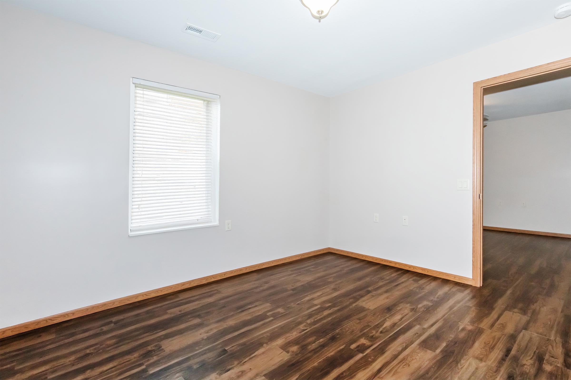 A vacant room with light beige walls and dark wooden flooring. There's a single window on the left with white blinds, and the room is well-lit. A doorway on the right leads to another space, suggesting a simple and modern design. The overall atmosphere is clean and uncluttered.