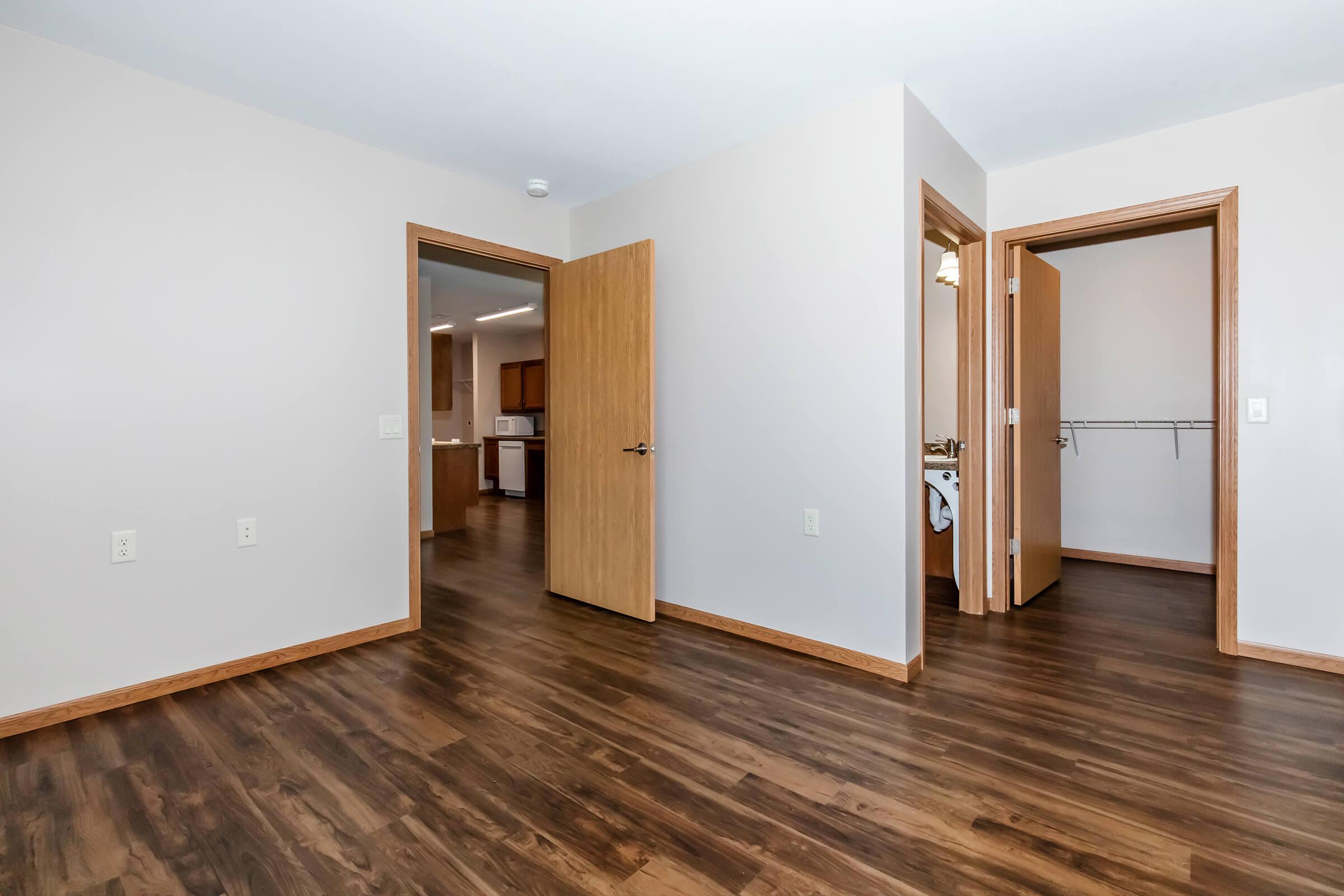 A spacious, empty room with light-colored walls and a wood floor. There are two open doors leading to adjacent rooms, one possibly a bathroom and the other a closet. Soft lighting highlights the clean and modern design of the space.