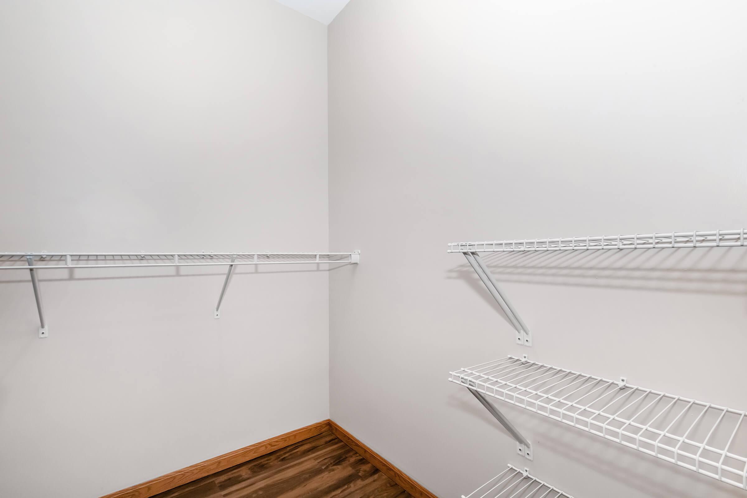 A clean, empty closet with white wire shelves attached to two walls, featuring a light beige wall and wooden flooring. The shelves are unoccupied, creating a spacious and organized appearance.
