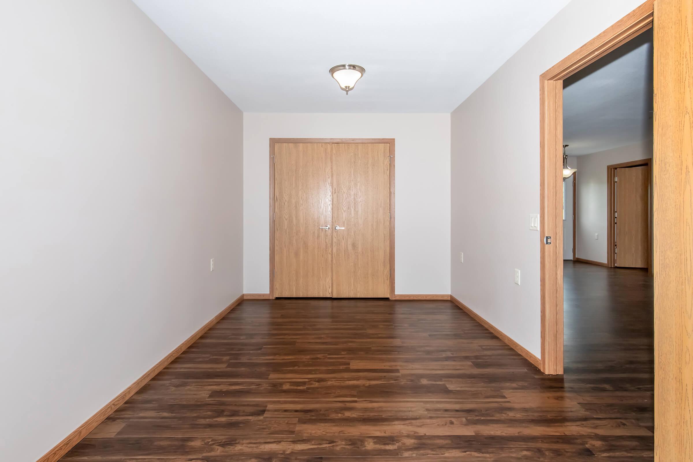 A spacious, well-lit hallway featuring hardwood flooring and light-colored walls. There are two wooden doors at the end leading to a closet or room, and another doorway on the right side that leads to a different area. A ceiling light fixture provides illumination.