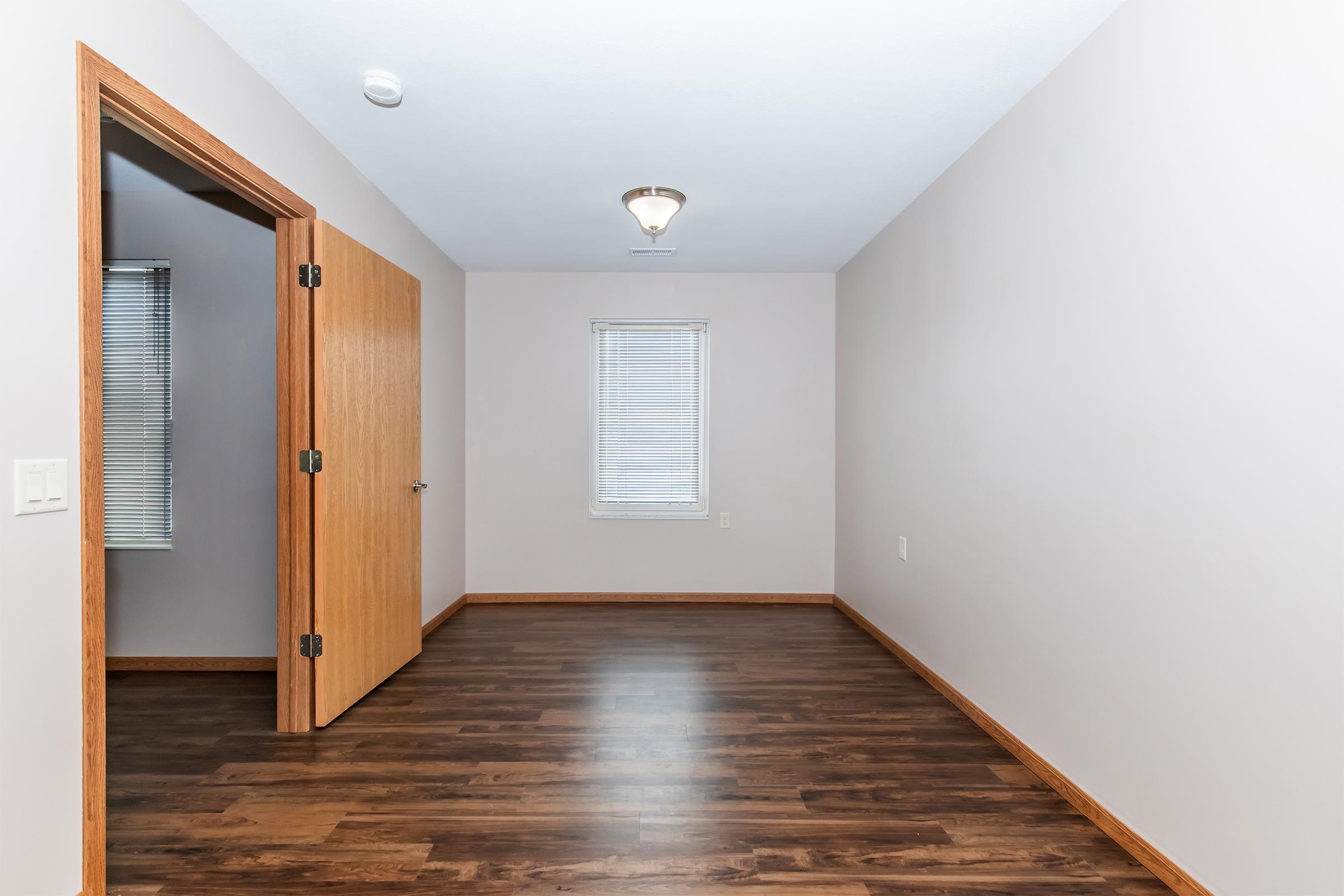 An empty room featuring light-colored walls, a wooden door, and a small window. The floor is dark wood, and the ceiling has a light fixture. The space is uncluttered and minimalistic, with natural light coming from the window.