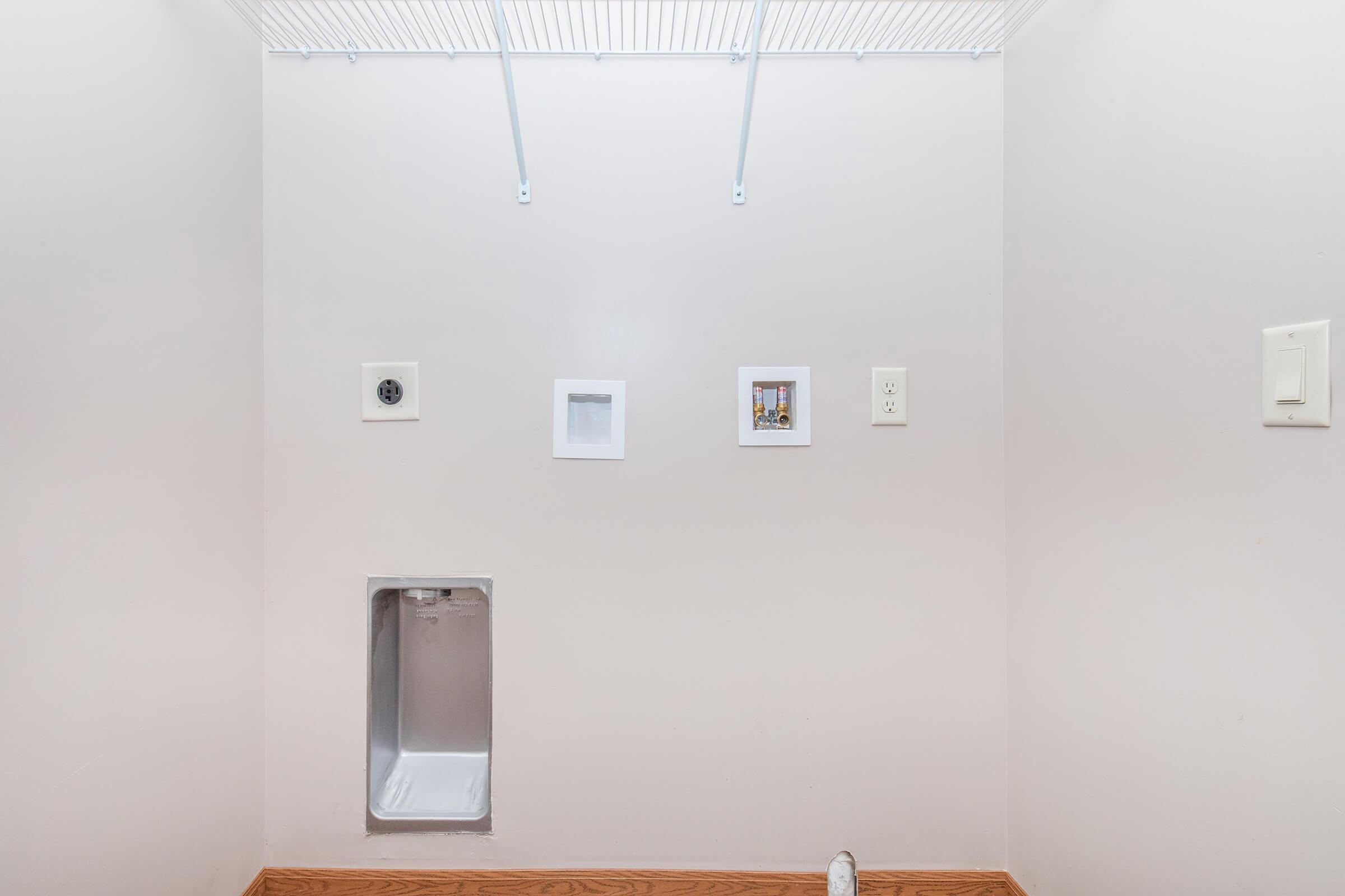 A spacious utility room with light-colored walls and wooden flooring. On the wall are three small, framed pictures and two electrical outlets. A pet door is visible at the bottom left, leading to the outside. The ceiling has a wire shelf for storage, and the overall space appears empty and clean.