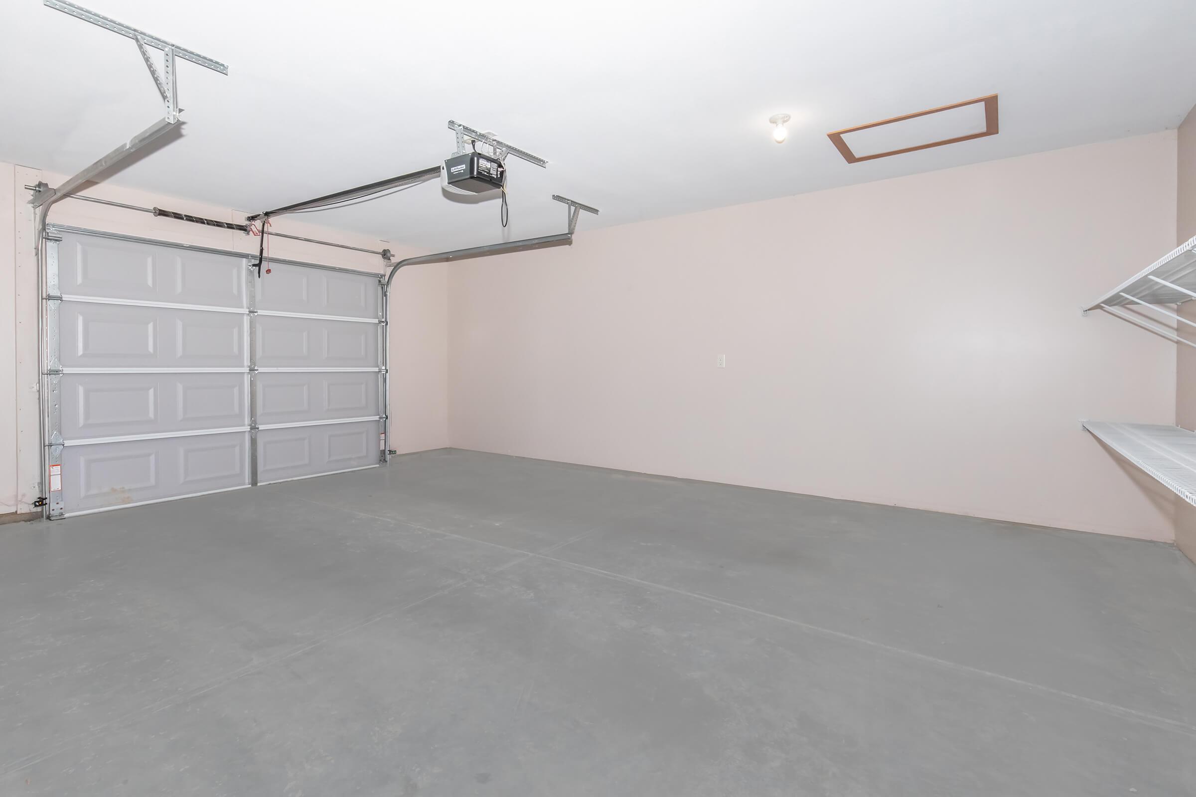 A clean, empty garage with a gray concrete floor and light pink walls. The garage features a double door with a metal frame, a ceiling light, and a small shelf on one side. There are no vehicles or clutter, creating a spacious and organized appearance.
