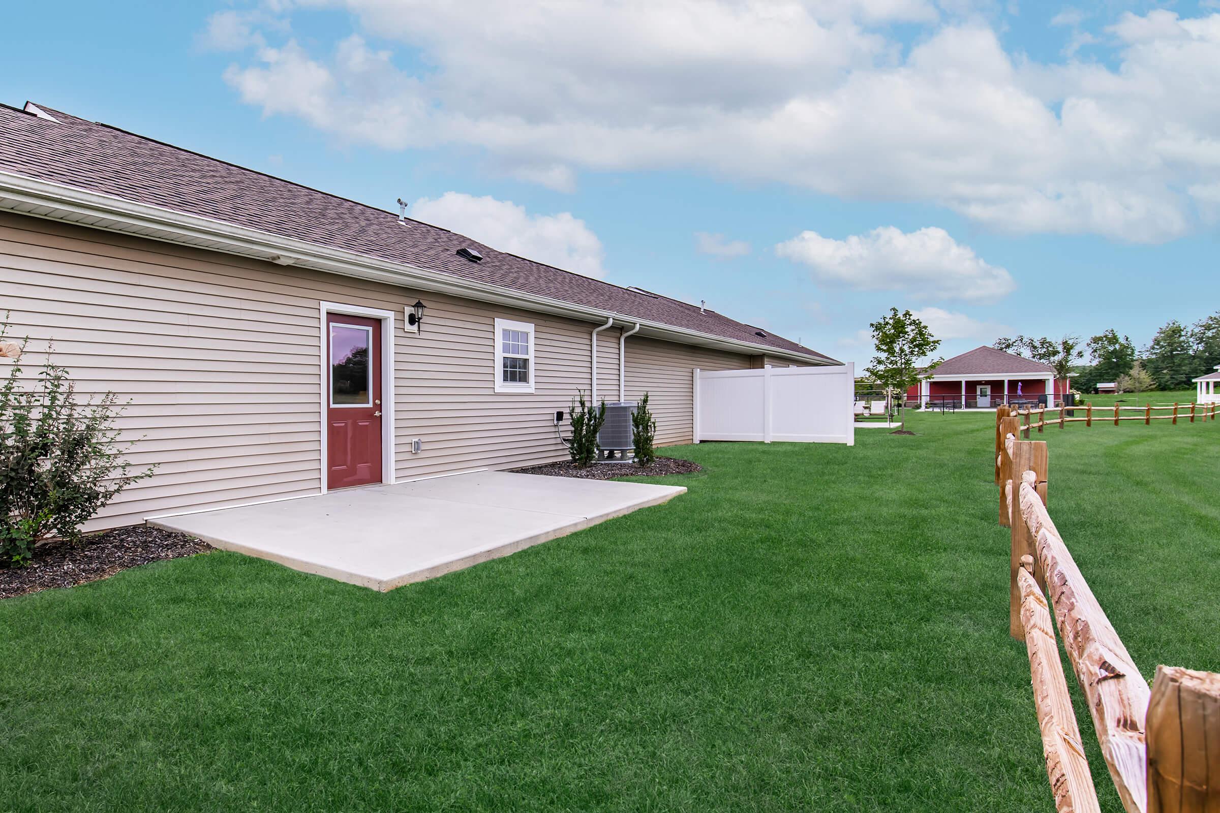 A view of the side yard of a residence featuring a well-maintained green lawn, a patio area with a door, and a fenced boundary. In the background, there is a red barn-like structure and a clear blue sky with scattered clouds.