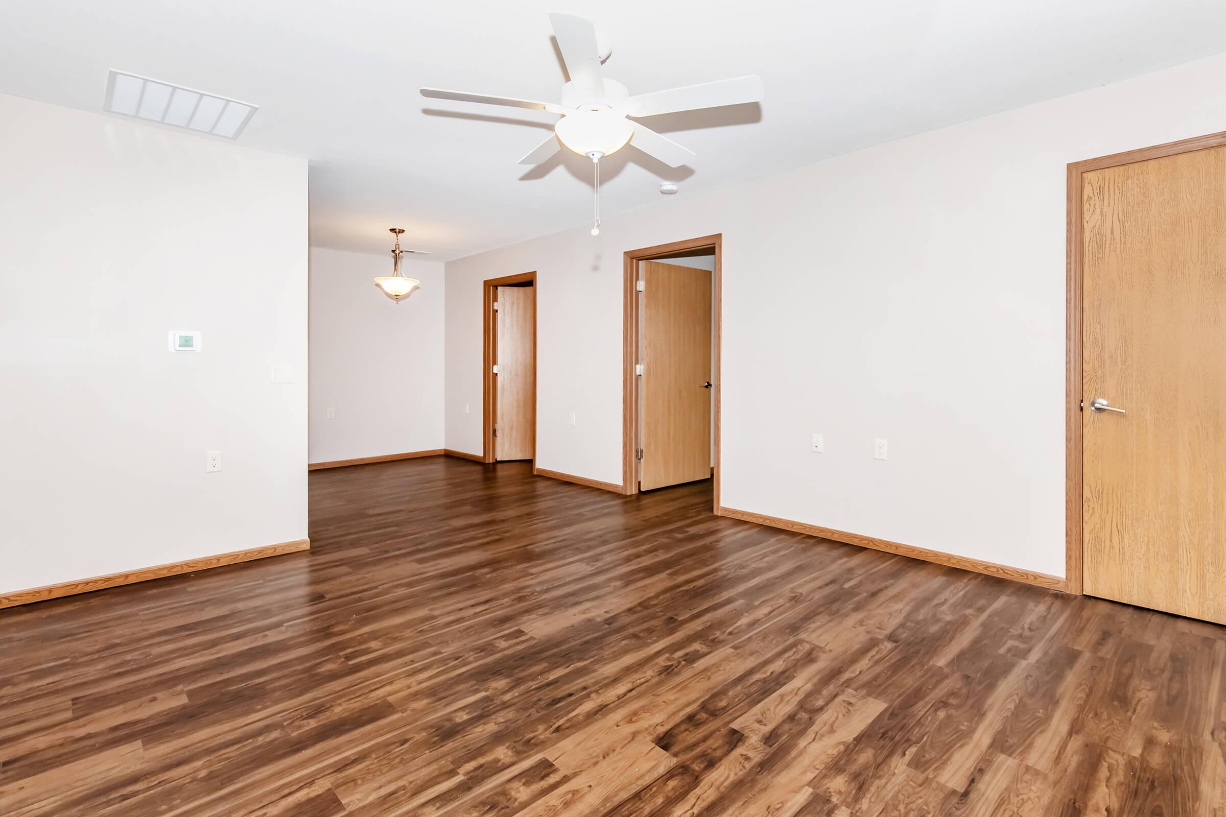 Spacious interior room featuring laminate wood flooring, a ceiling fan, and neutral-colored walls. The room has multiple doorways leading to other spaces, with a warm light fixture hanging from the ceiling, creating a welcoming atmosphere.