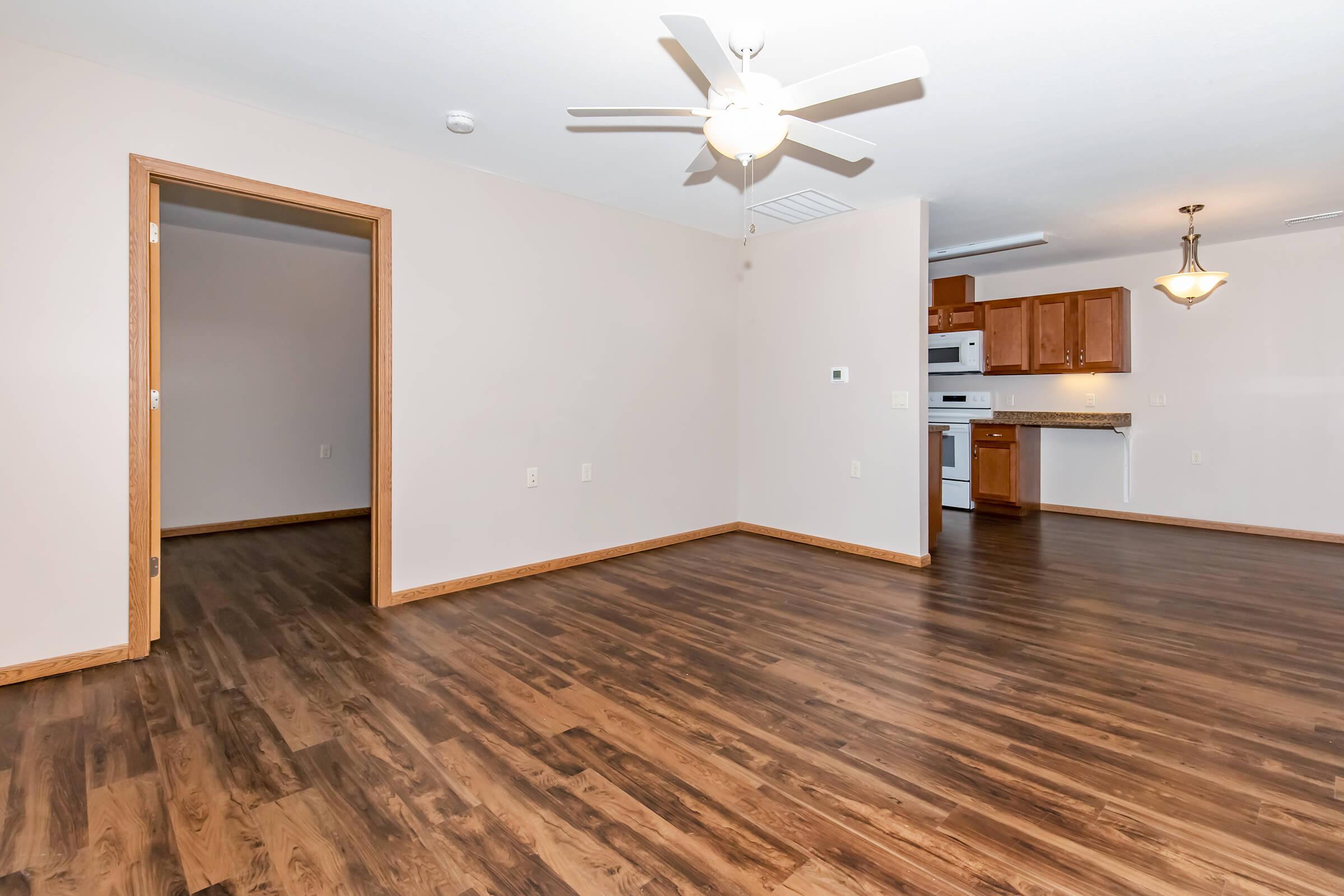 Spacious, light-filled living area featuring wood-like flooring, a ceiling fan, and an adjoining kitchen with wooden cabinets and a range. A doorway leads to another room, enhancing the open layout. The walls are painted in a neutral tone, creating a warm and inviting atmosphere.