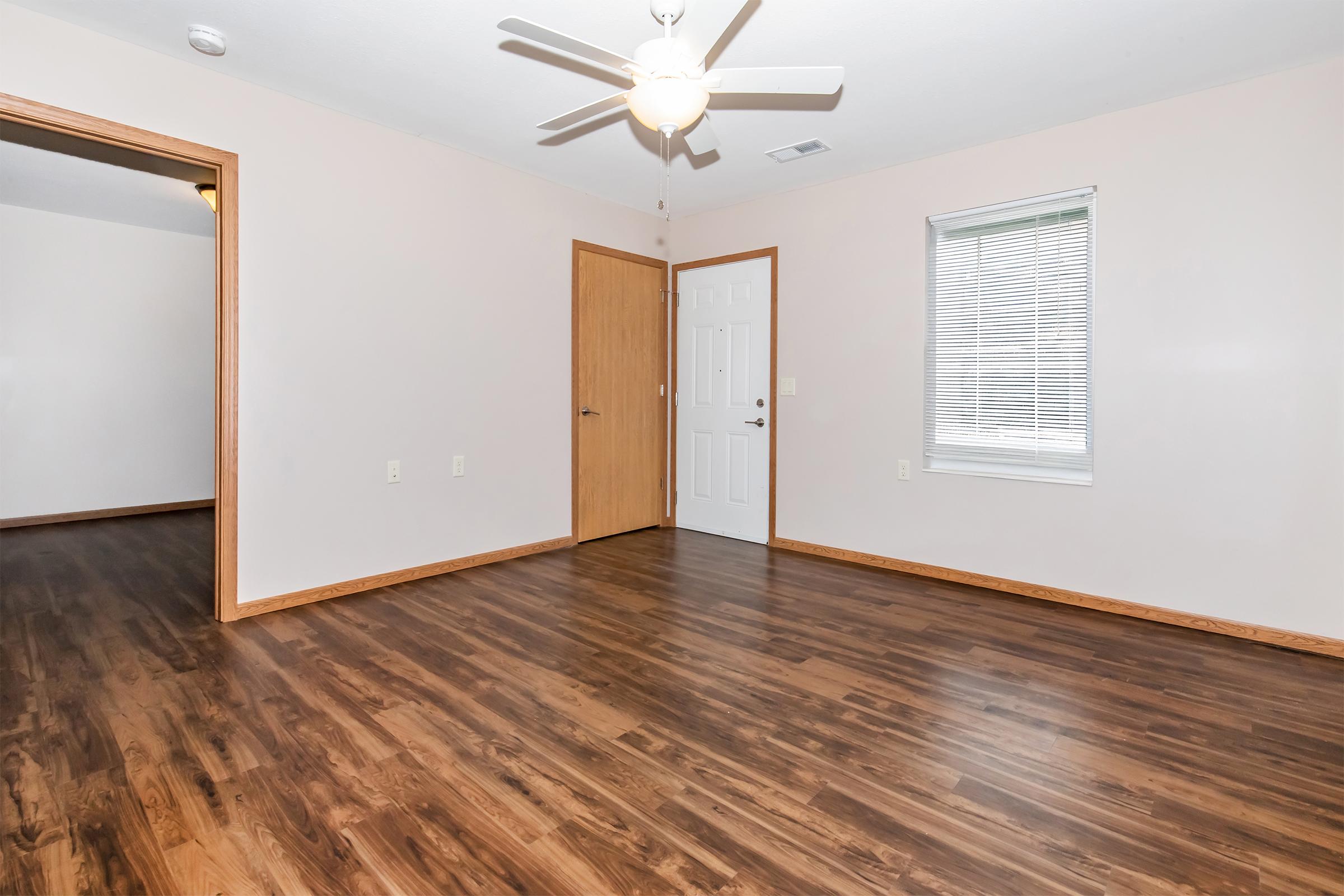 A spacious room featuring light brown walls and dark wooden flooring. It has a ceiling fan, a door leading to another room, and a door leading outside. A small window with blinds allows natural light to enter. The room is empty and ready for furnishing.