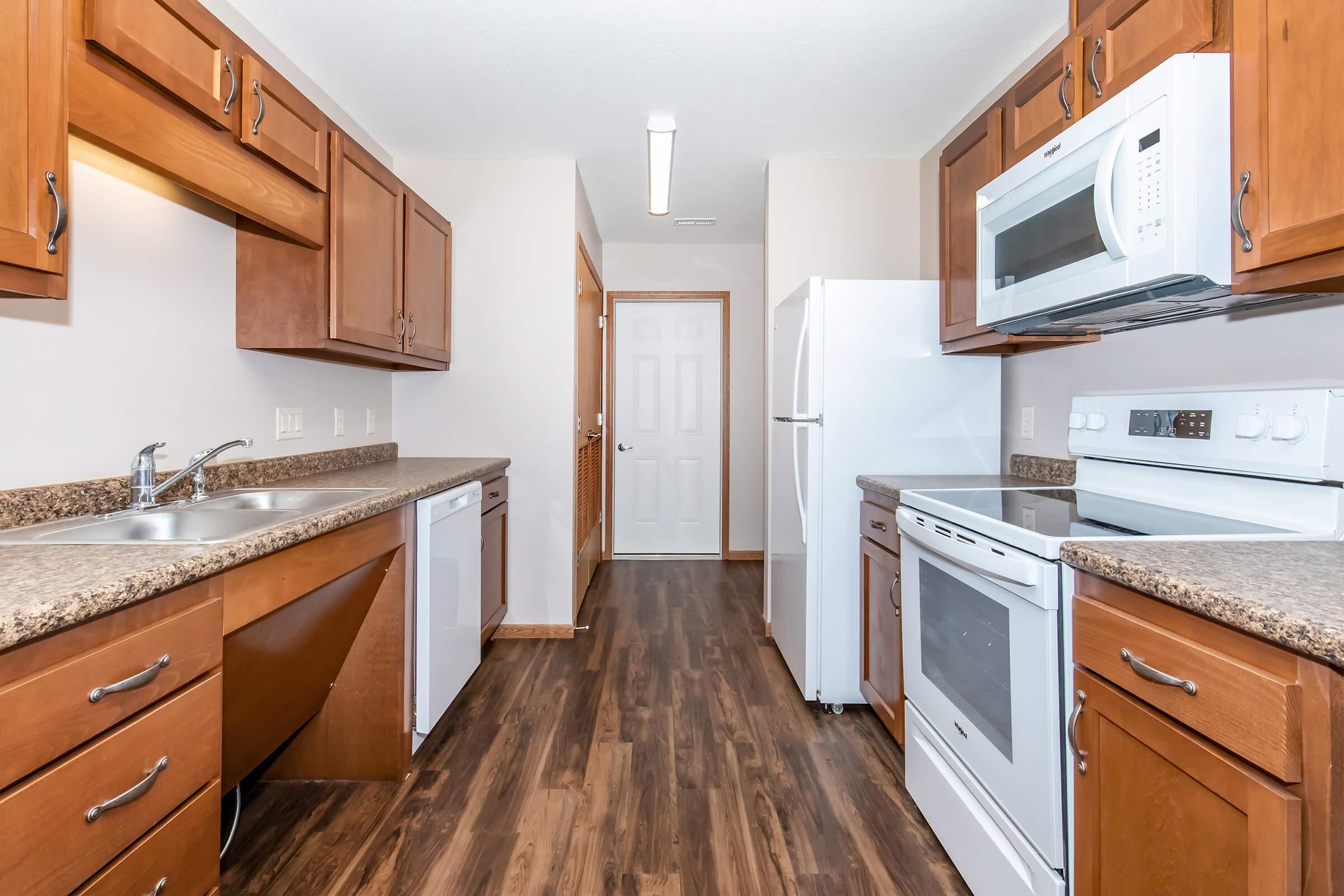 Modern kitchen featuring wooden cabinets, granite countertops, and stainless steel appliances, including a microwave, oven, and dishwasher. The space is well-lit with overhead lighting and has a door leading to another room. The flooring is a rich wood finish, enhancing the warm atmosphere of the kitchen.